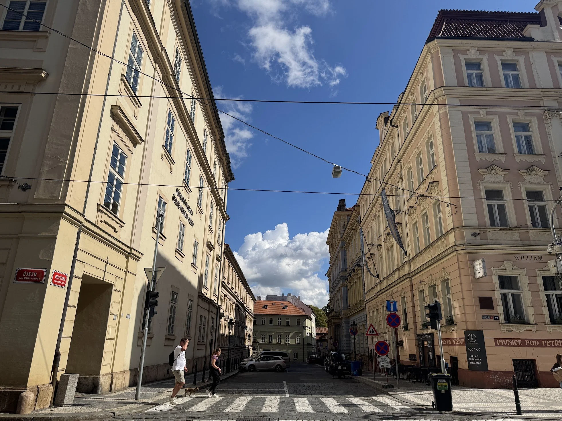 City street scene with tall, ornate buildings on both sides, people crossing the street, parked cars, and signposts with traffic and parking symbols. The sky is blue with some clouds.