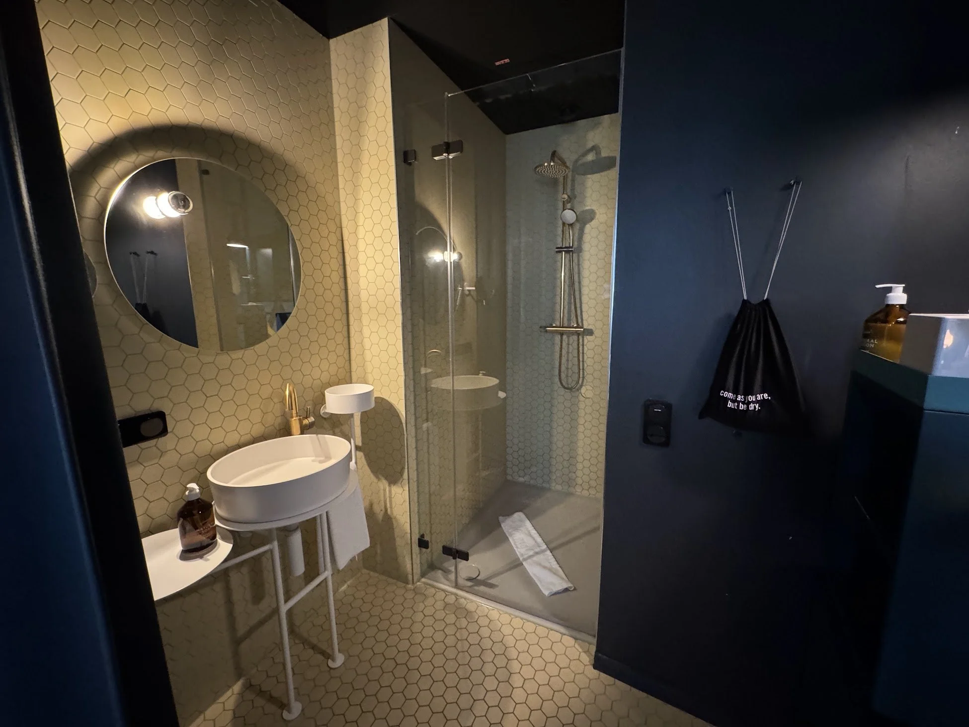 Bathroom with a round mirror, yellow hexagon tile wall, small white sink, black soap dispenser, glass shower enclosure with shower head and controls, dark walls, hanging black bag, and amber soap bottle on shelf.