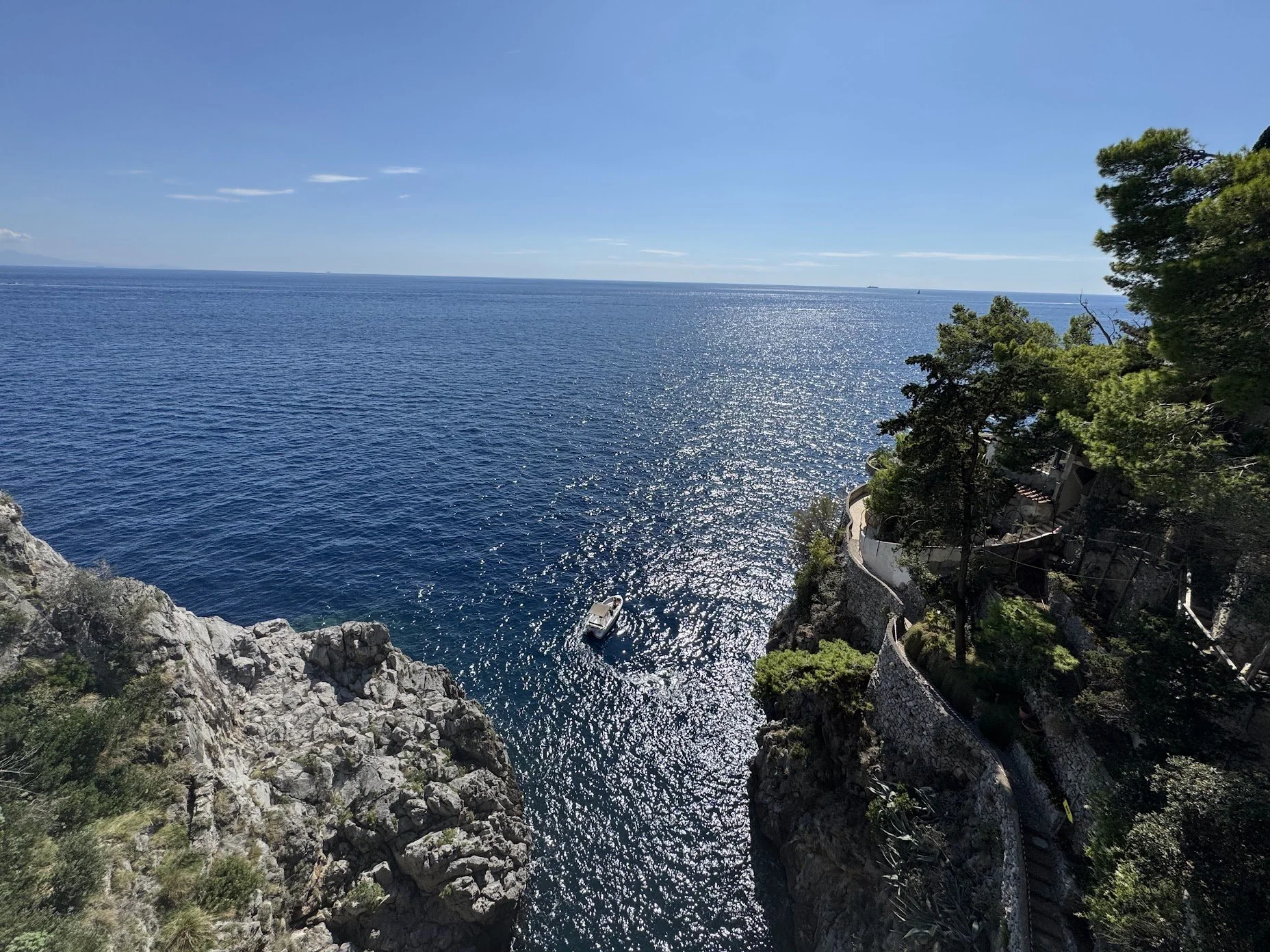 A panoramic view of the sea with calm blue water reflecting sunlight, a small boat floating near rocky cliffs, and a coastline with greenery and trees on the right side under a clear blue sky.