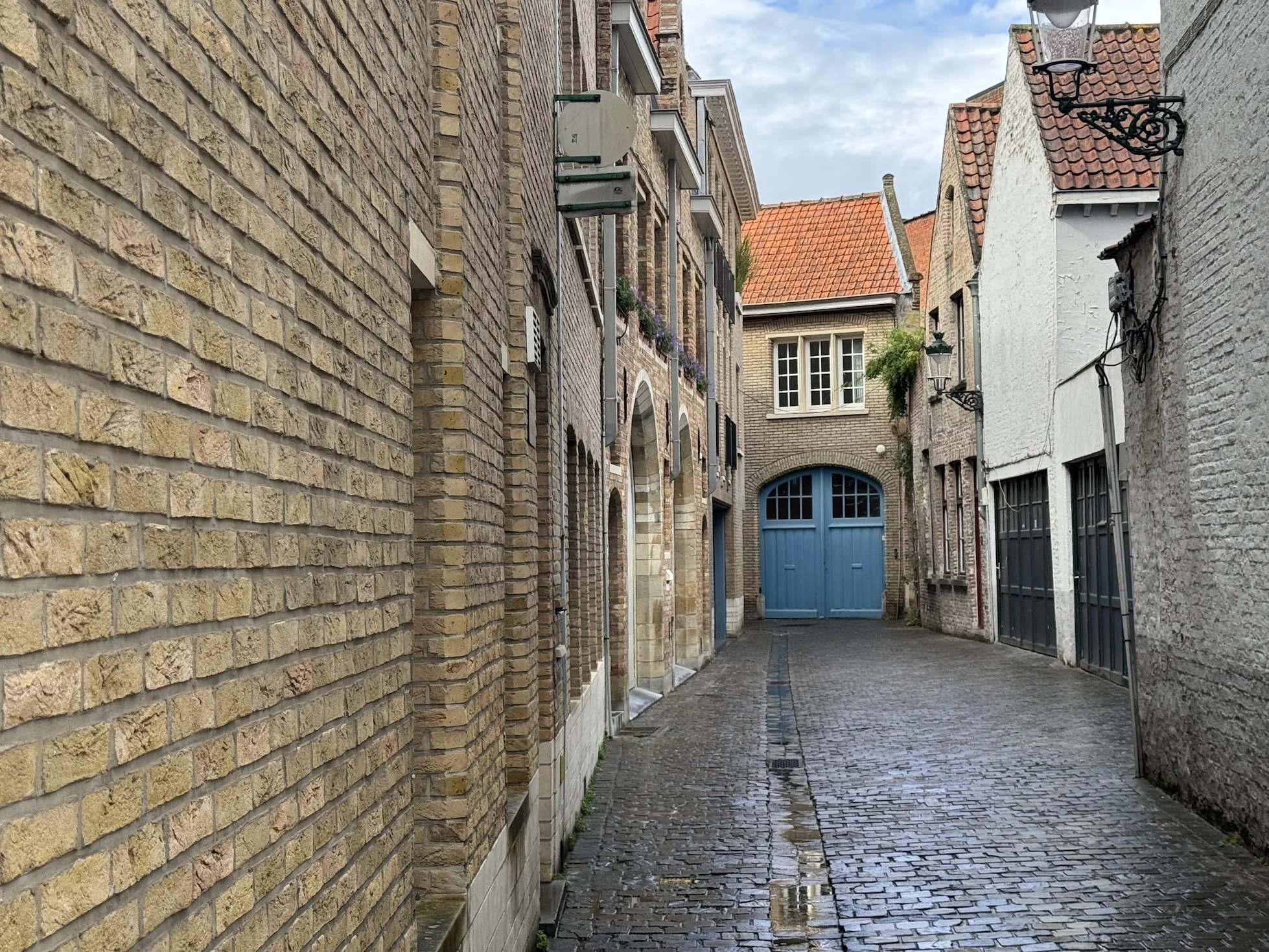 A narrow cobblestone alley surrounded by old brick buildings with windows and plants, leading to a blue door under an archway, with a partly cloudy sky above.