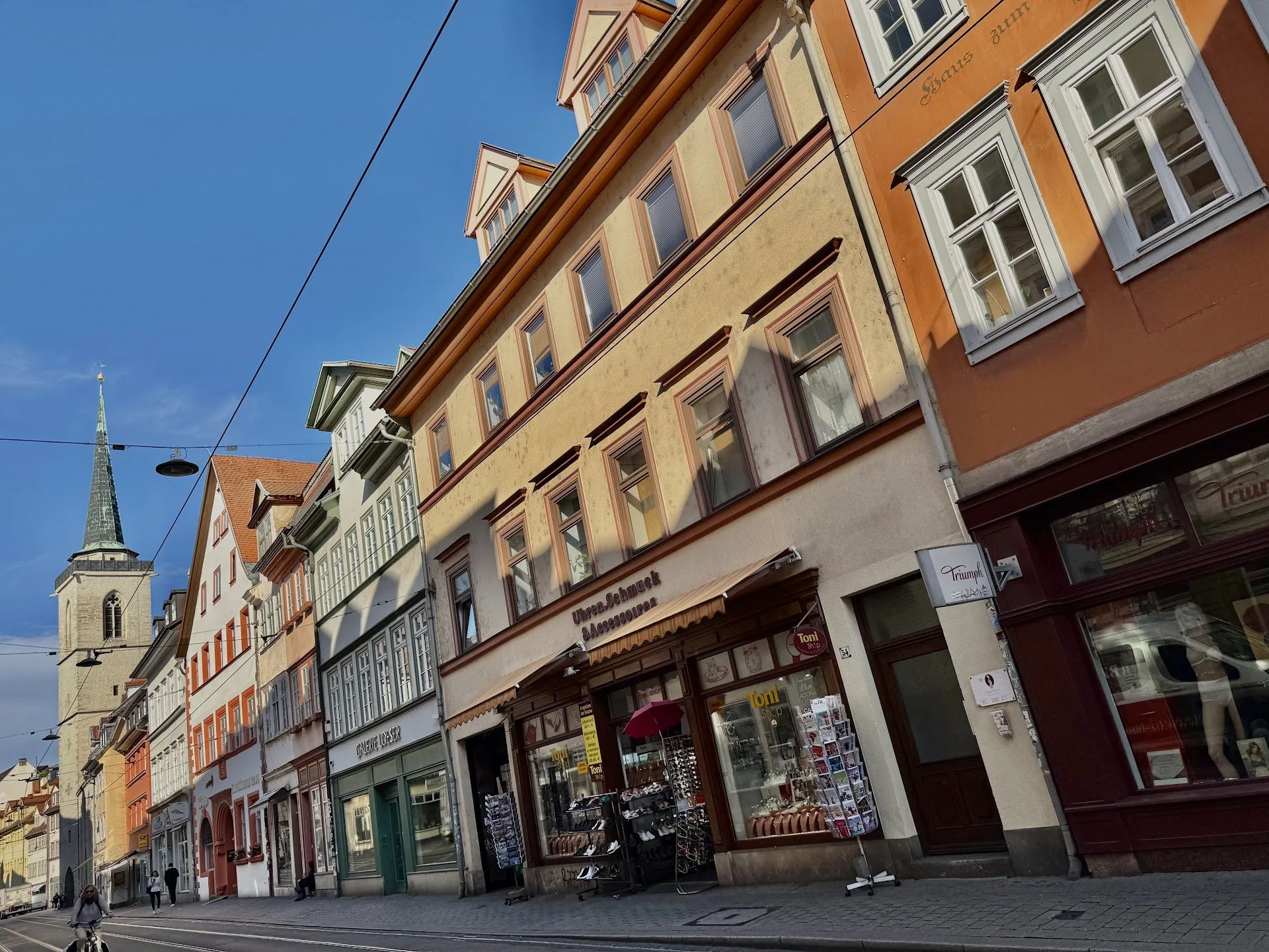 View of colorful multi-story buildings on a city street with tram tracks, awnings, and a church steeple in the background under a clear blue sky.