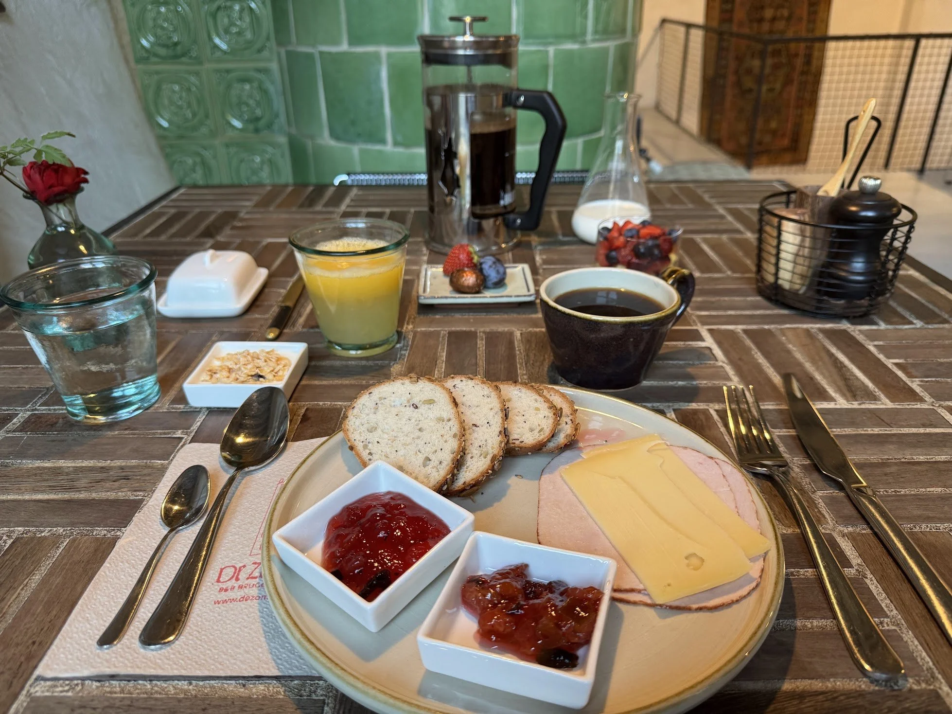 A breakfast spread on a dining table featuring a plate of sliced bread, slices of cheese and ham, and three small bowls filled with strawberry, blueberry, and mixed fruit jam. There is a cup of black coffee, a glass of orange juice, a glass of water,