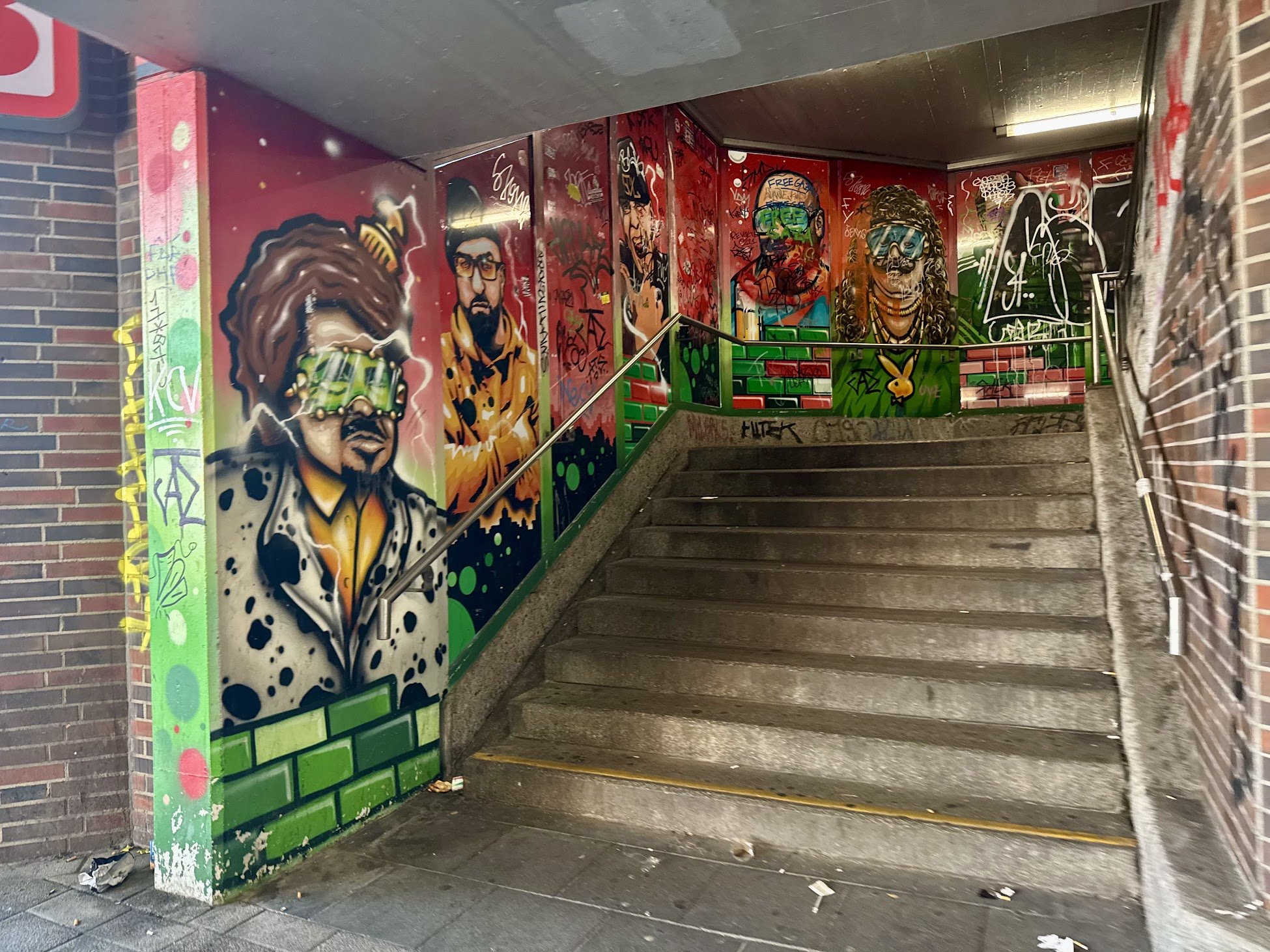 Graffiti artwork on a staircase wall depicting various stylized portraits of people with vibrant colors, spray paint tags, and abstract designs in an urban setting.