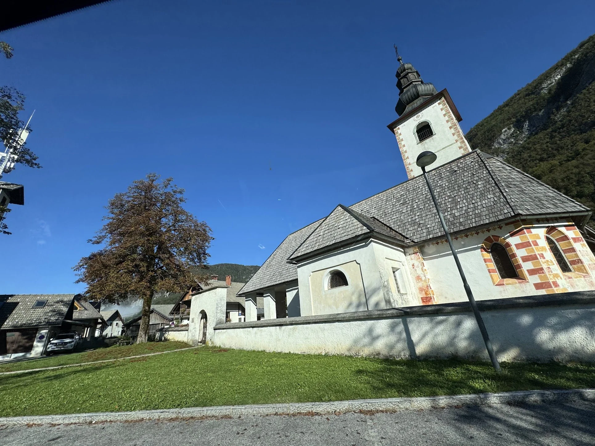A white church with a tall steeple and a clock on its front, set against a bright blue sky with few clouds. There are trees and houses around the church, with a grassy lawn in the foreground and a parking area visible on the left.
