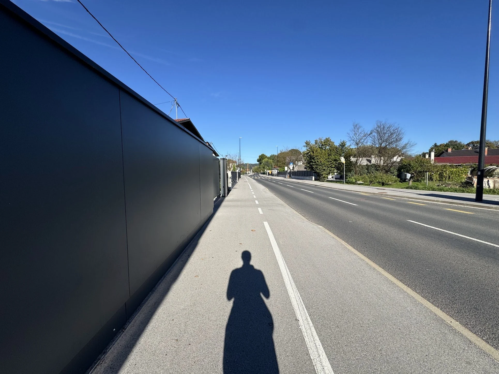 Empty sidewalk beside a street with a black wall on the left and trees with some houses in the background. The shadow of a person and their phone are cast on the sidewalk, indicating the photo was taken by the person.