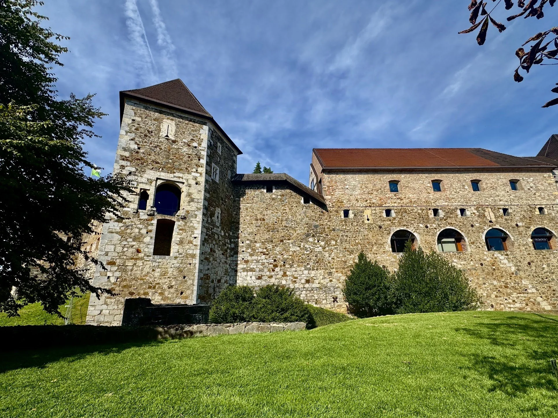 A historic stone building with a tower, small windows, and a red-tiled roof, surrounded by green trees and grass, under a partly cloudy blue sky.