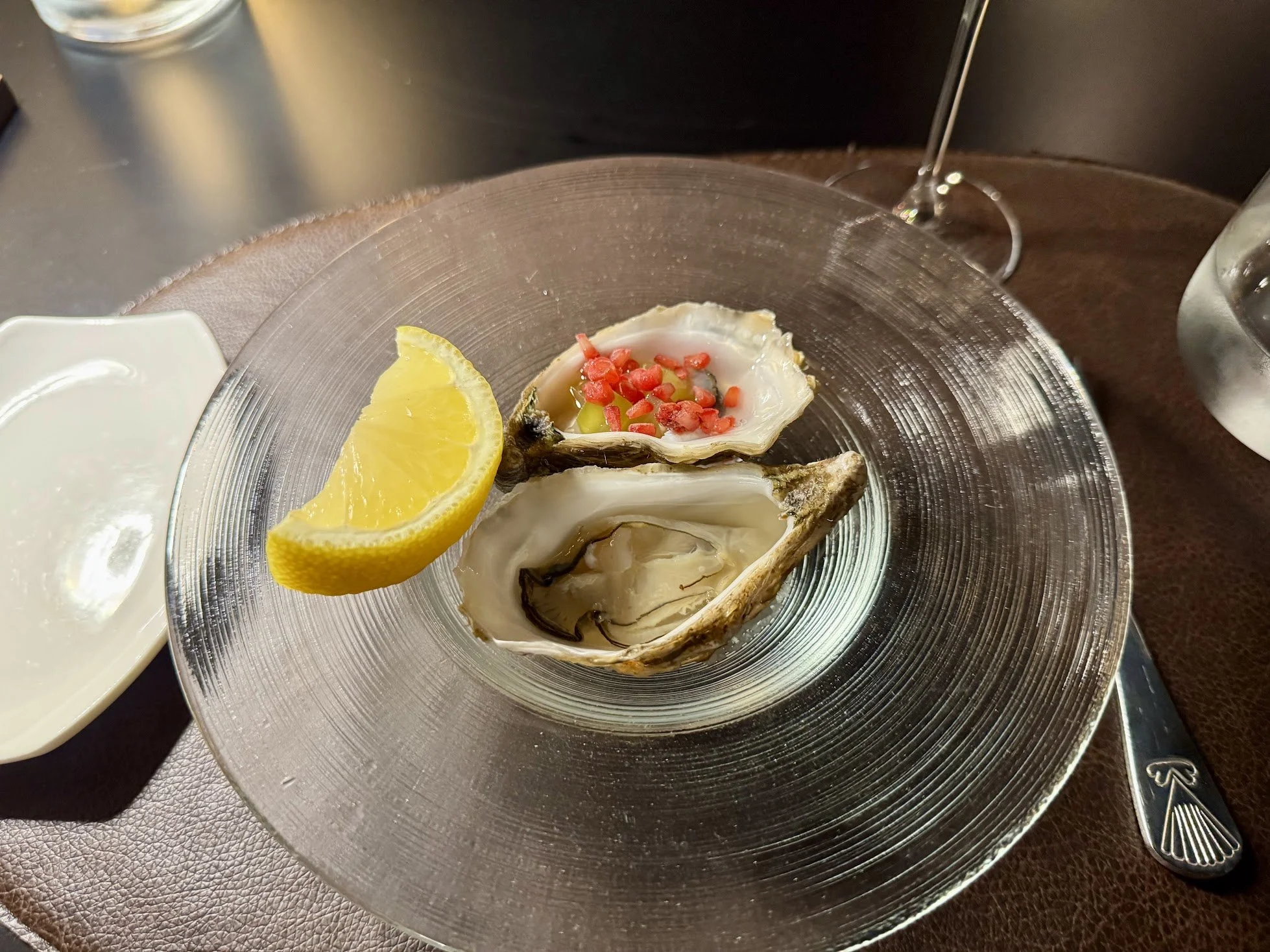 A glass plate with a fresh oyster topped with red chopped tomatoes or peppers, and a lemon wedge beside it. There are plates and utensils on a dark table.