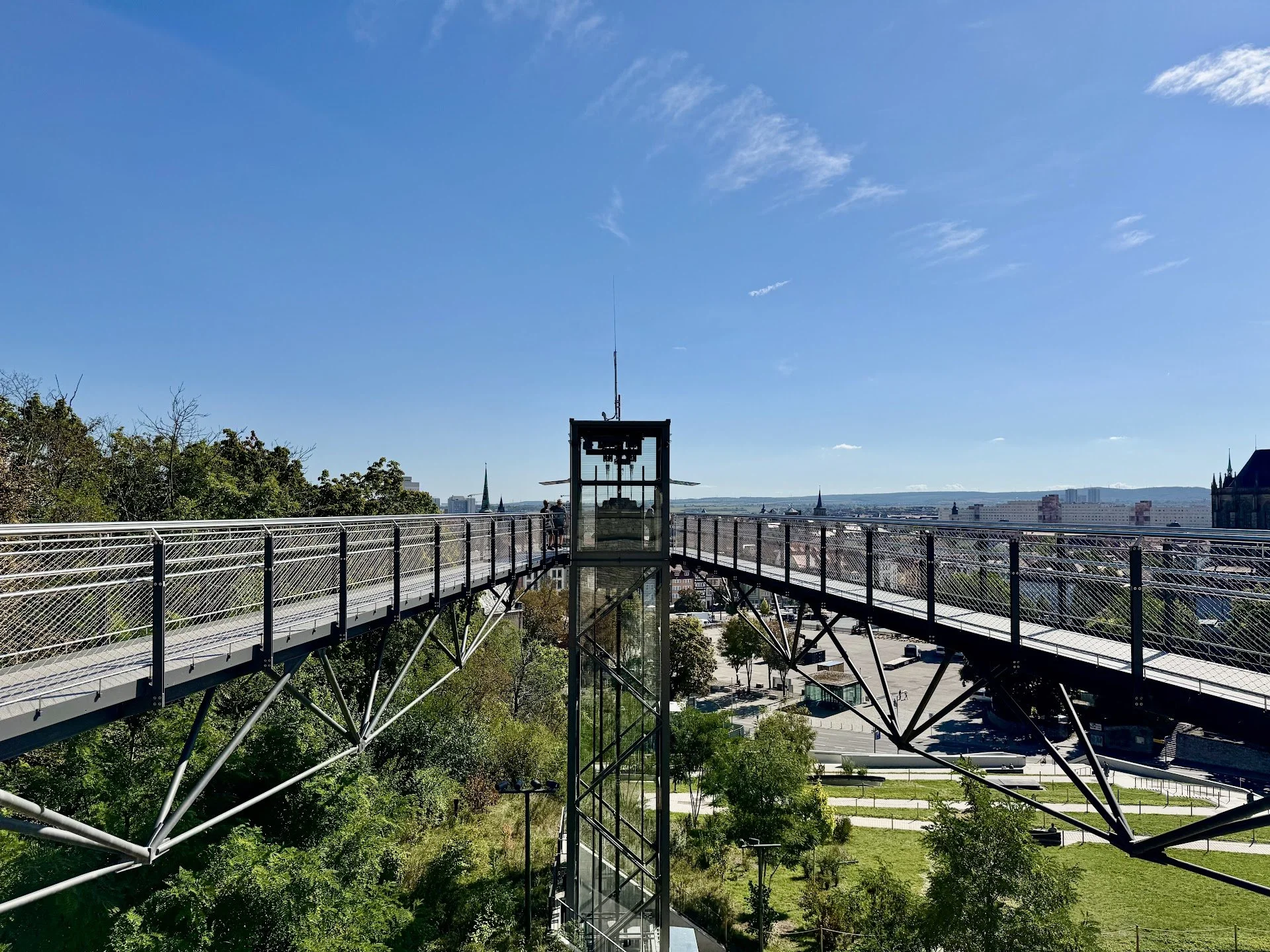 View from high above of a modern glass elevator and a steel suspension walkway with a cityscape and blue sky in the background.