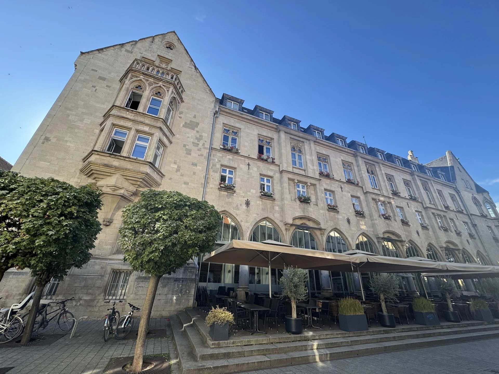 A historic multi-story stone building with arched windows and decorative architectural details, featuring a corner turret, a rooftop attic with dormer windows, and a ground-level outdoor patio with umbrellas, potted plants, and bicycles parked nearby