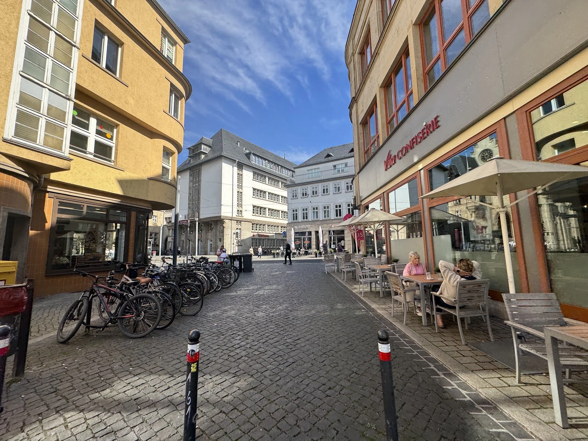 An outdoor scene with people sitting at tables in front of a Cafe. There are bicycles parked on a cobblestone street. Several buildings with large windows and signs are visible in the background. People are walking or sitting, enjoying a sunny day.