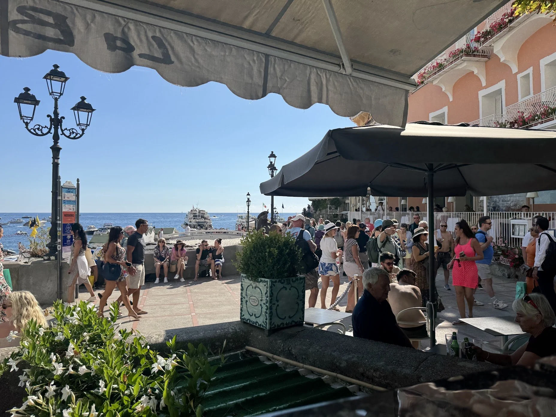 Crowded outdoor seaside café scene with people walking and sitting, bordered by buildings with balconies and flowers, and a view of the ocean with boats and a yacht.