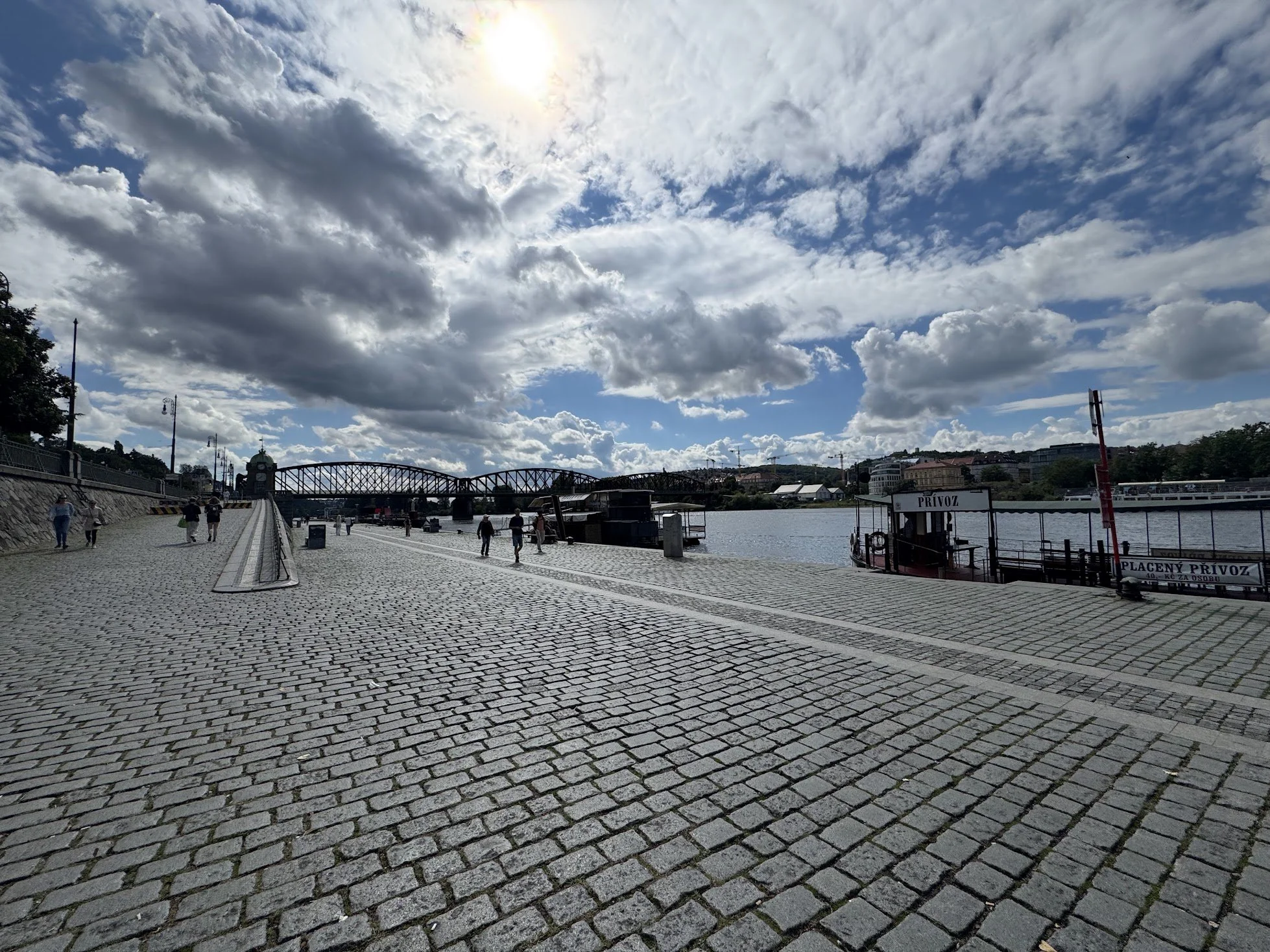 A riverside walkway with cobblestone pavement, people walking, a bridge over the river, and a partly cloudy sky with the sun shining.