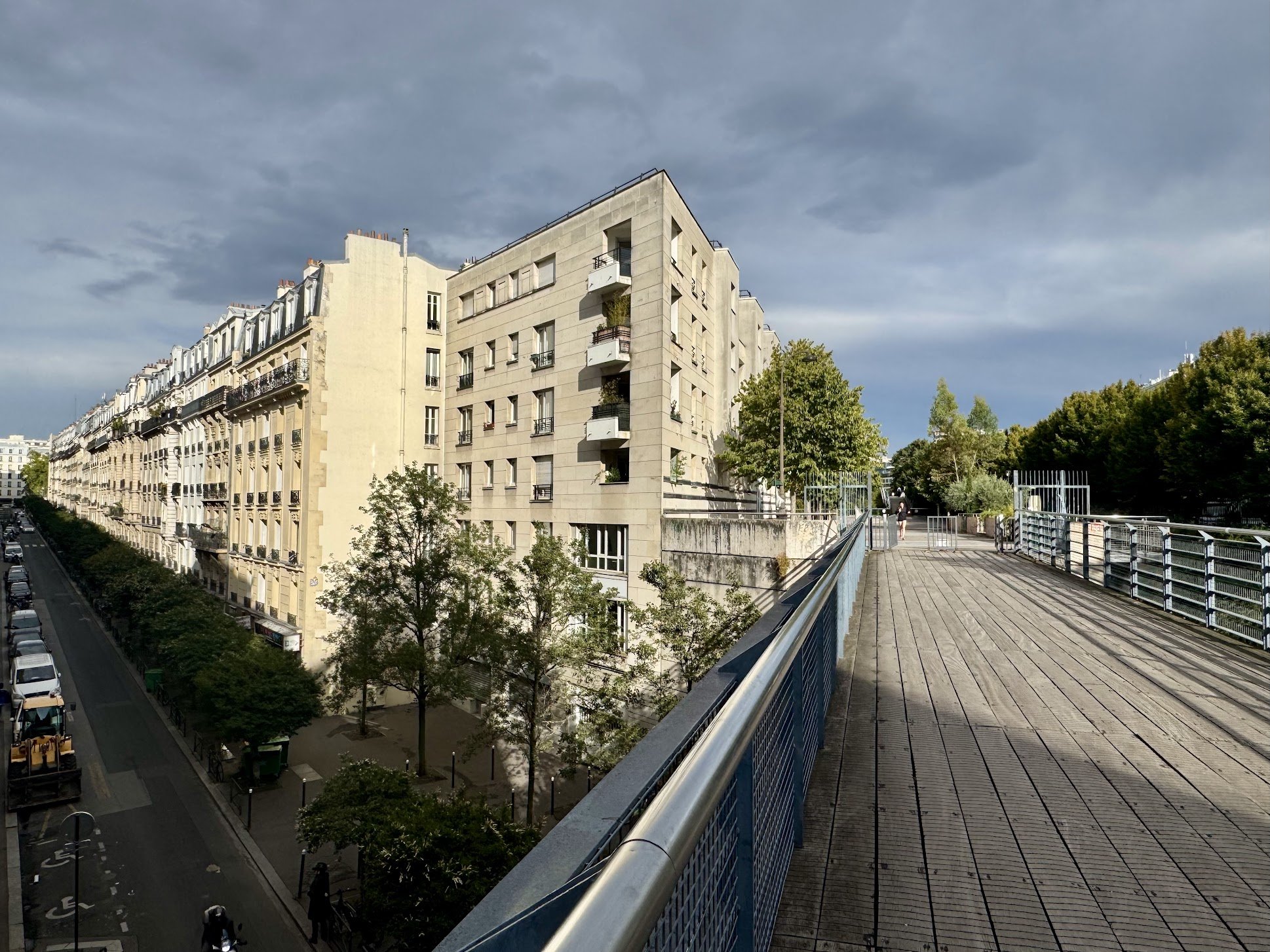 View of a multi-story beige residential building with balconies, trees lining the street below, and a blue sky with scattered clouds. A pedestrian walkway with a metal railing is in the foreground.