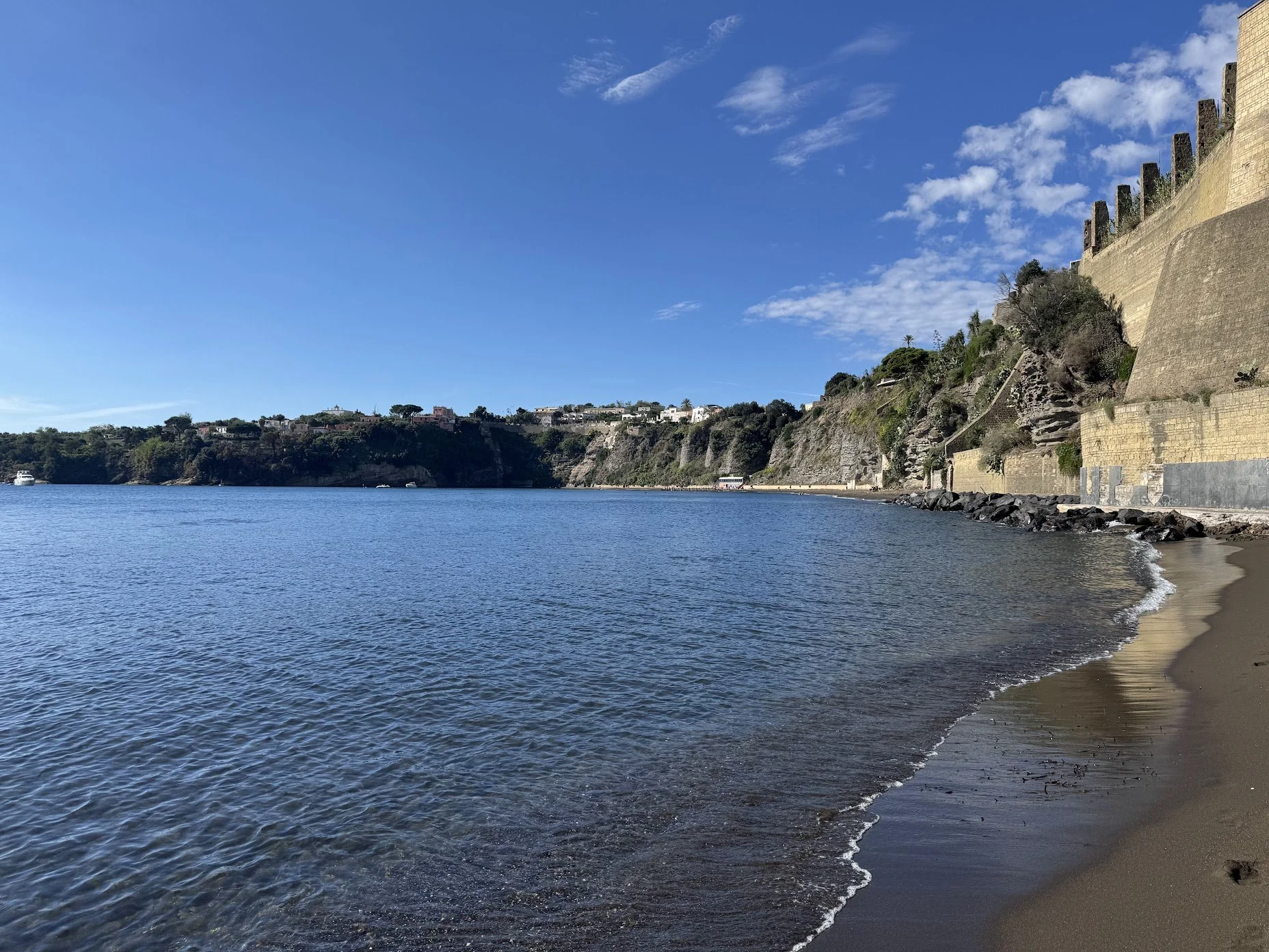 A beach with a sandy shoreline and calm water, bordered by a historic stone wall on the right and houses on a hill in the background under a clear blue sky with a few clouds.