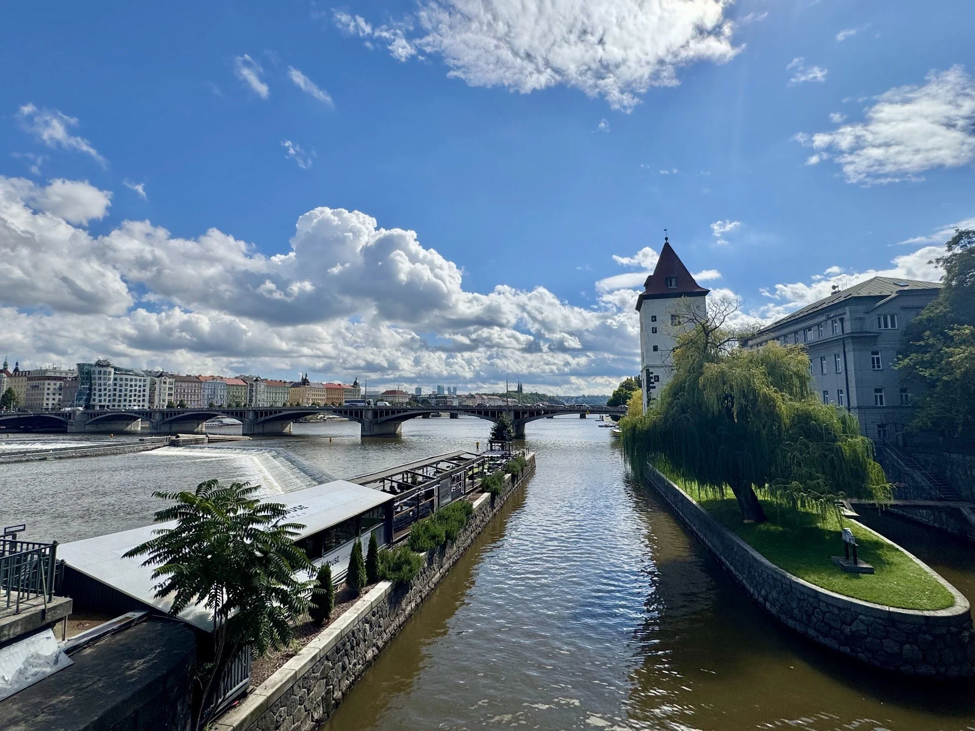 View of a city river with buildings and a bridge in the background, a tower with a pointed roof, and a partly cloudy sky.