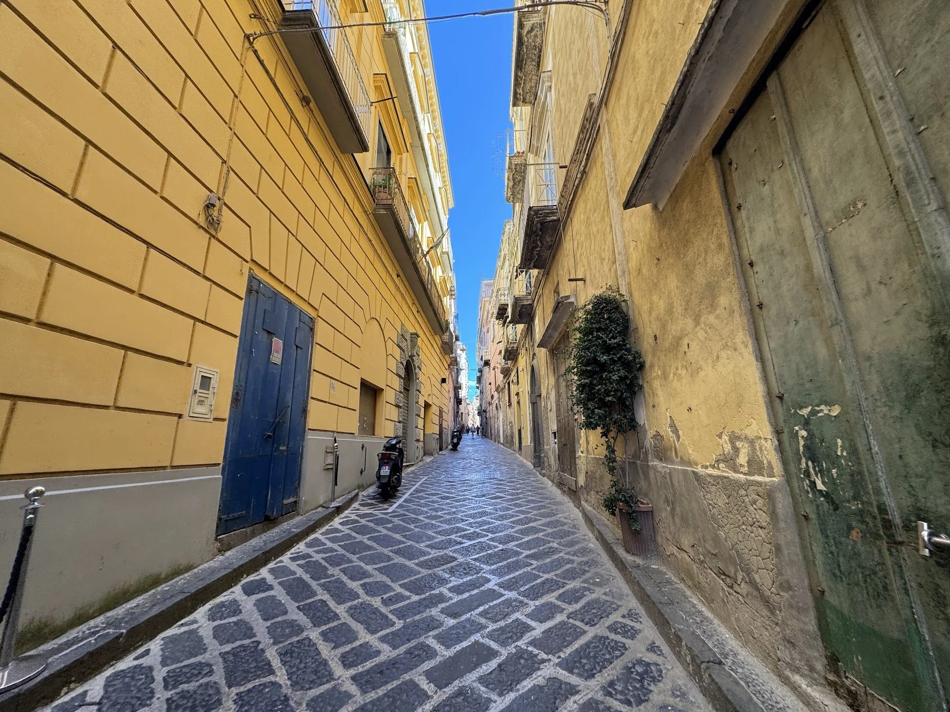 A narrow cobblestone street in an old European city lined with yellow and weathered beige buildings, some with balconies and plants, under a clear blue sky.