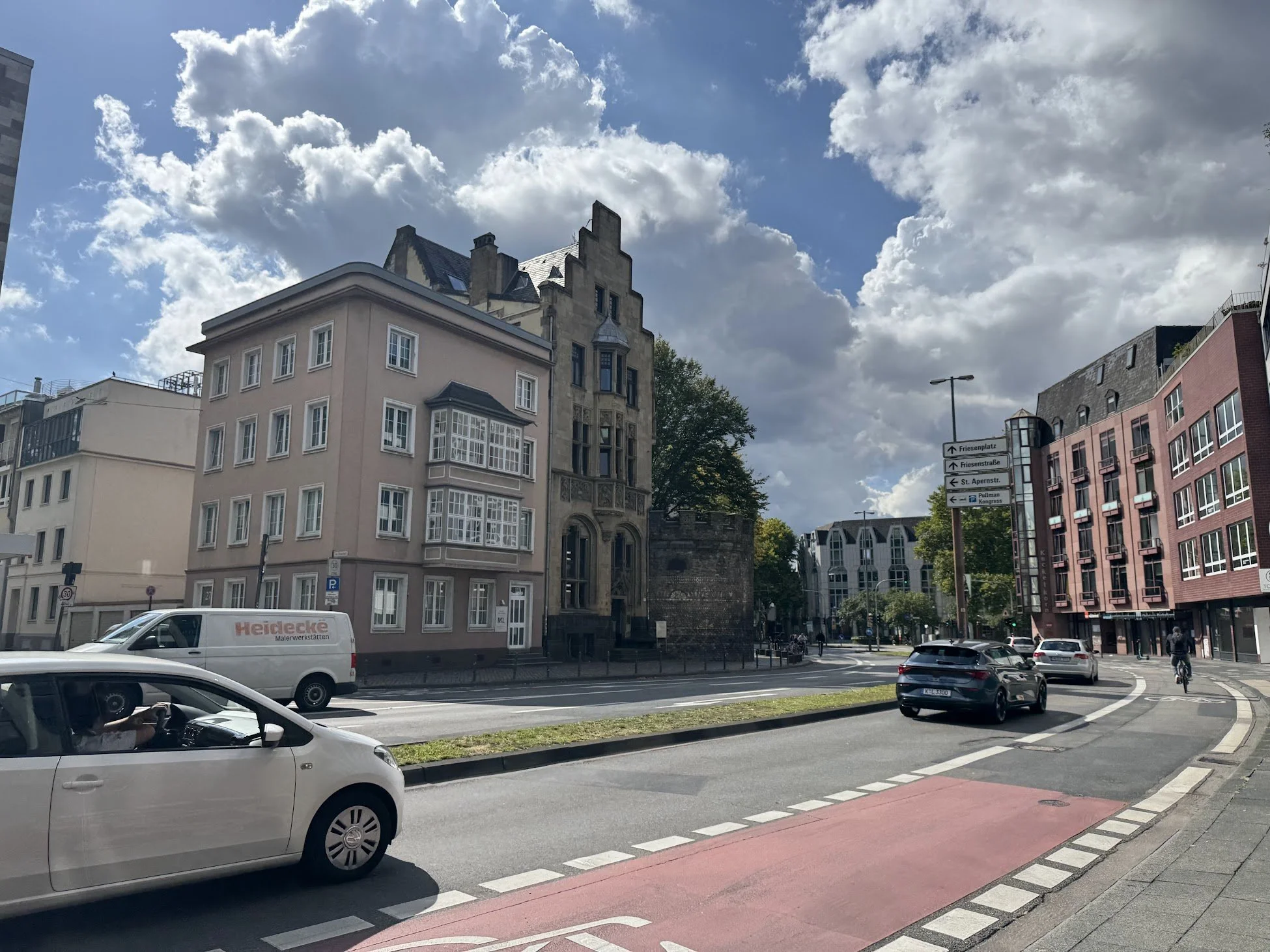 City street with modern buildings, cars on the road, signage indicating directions, and a partly cloudy sky.