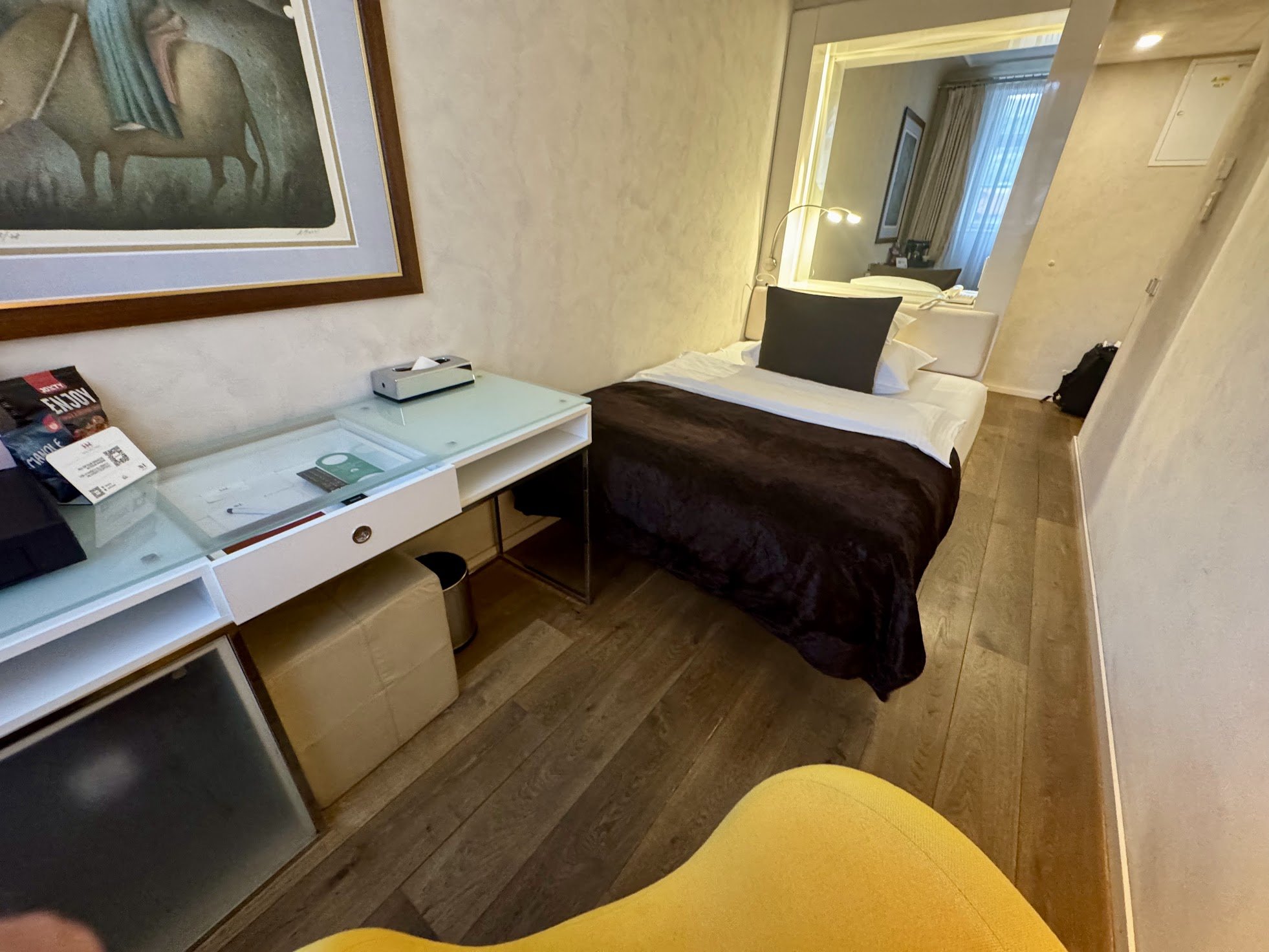 A hotel room features a bed with white linens and dark brown blankets, a nightstand with a lamp, a framed artwork hanging above the bed, and a window with sheer curtains. The room has light-colored wallpaper and wooden flooring.