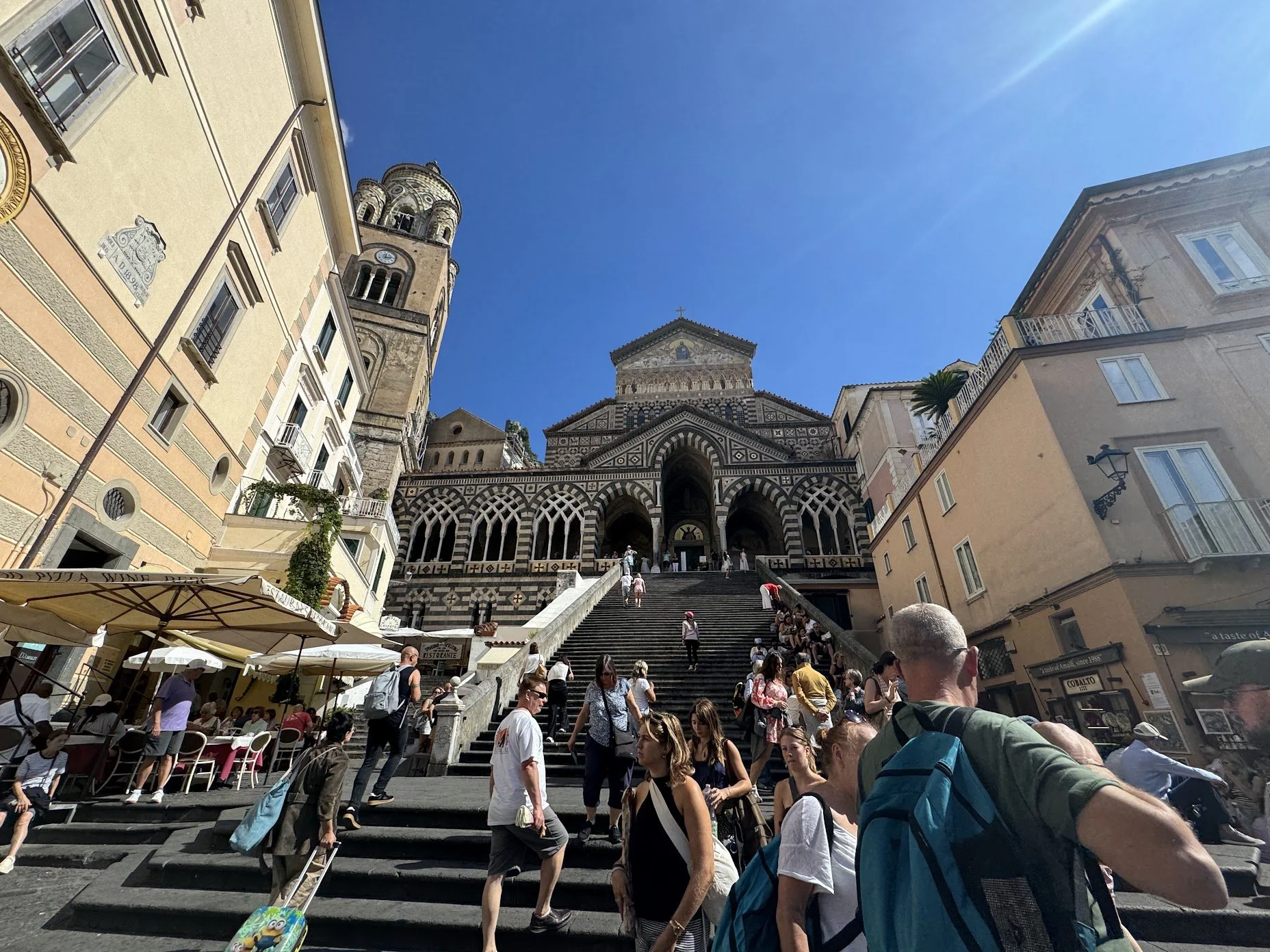Crowded staircase leading up to a historic church with a patterned facade and a tall bell tower, surrounded by city buildings, people, and outdoor cafe seating under umbrellas on a sunny day.