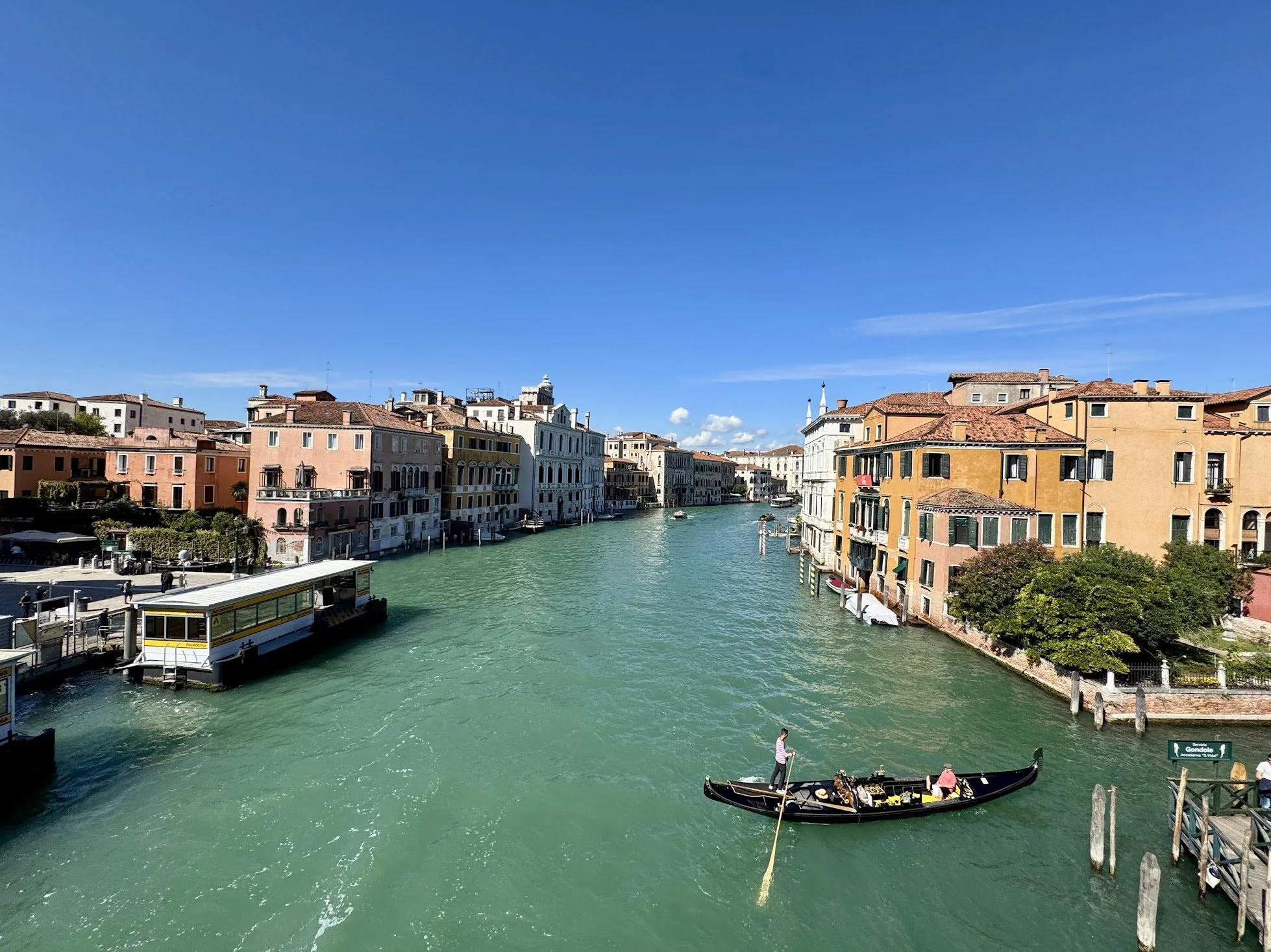 Colorful buildings on either side of a canal in Venice, Italy, with a gondola and a water bus navigating the waterway under a clear blue sky.