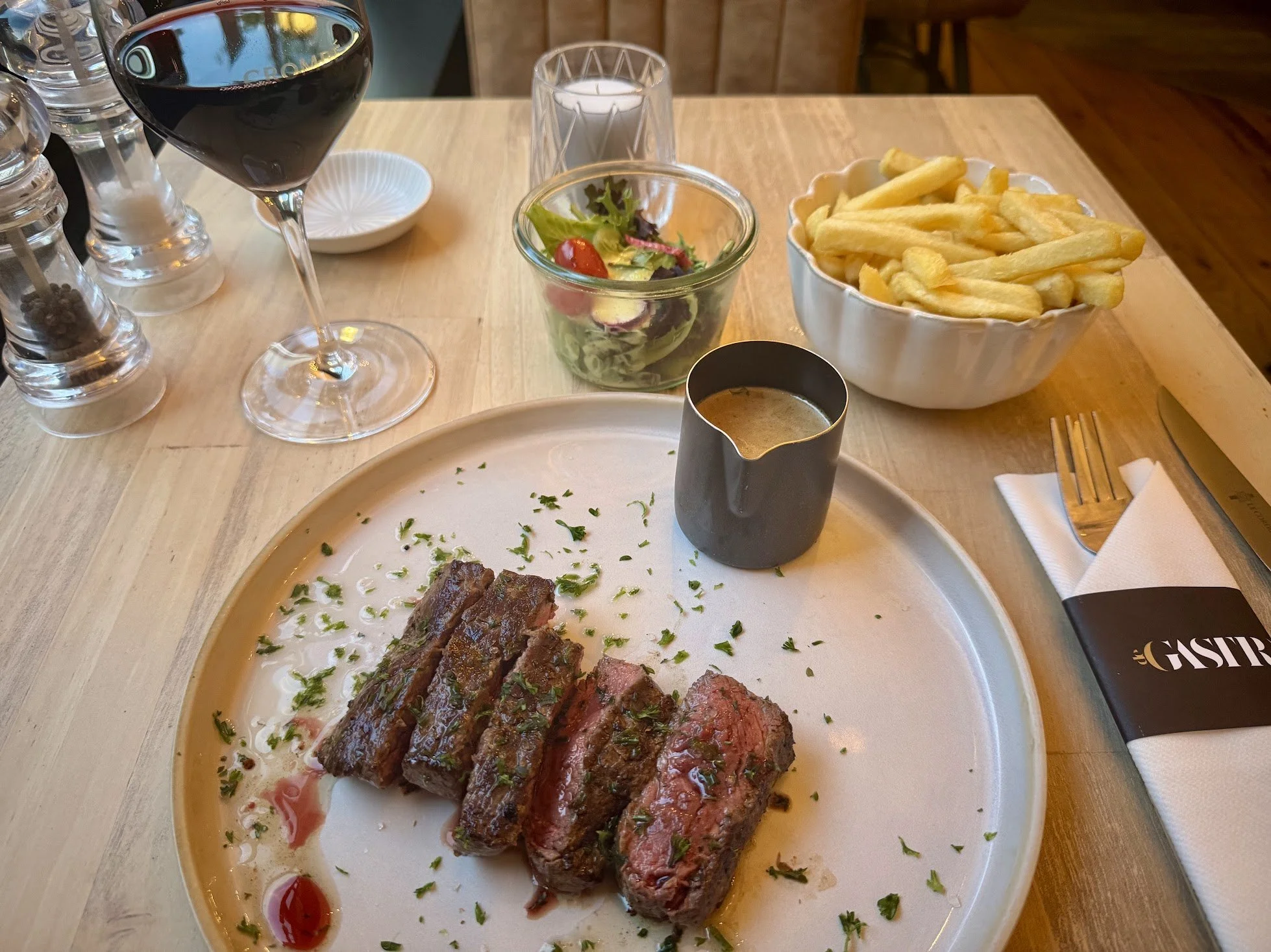 A restaurant table with a glass of red wine, a small salad, a bowl of French fries, a plate with cooked steak slices topped with herbs, a small pitcher of sauce, some condiments, and a candle.