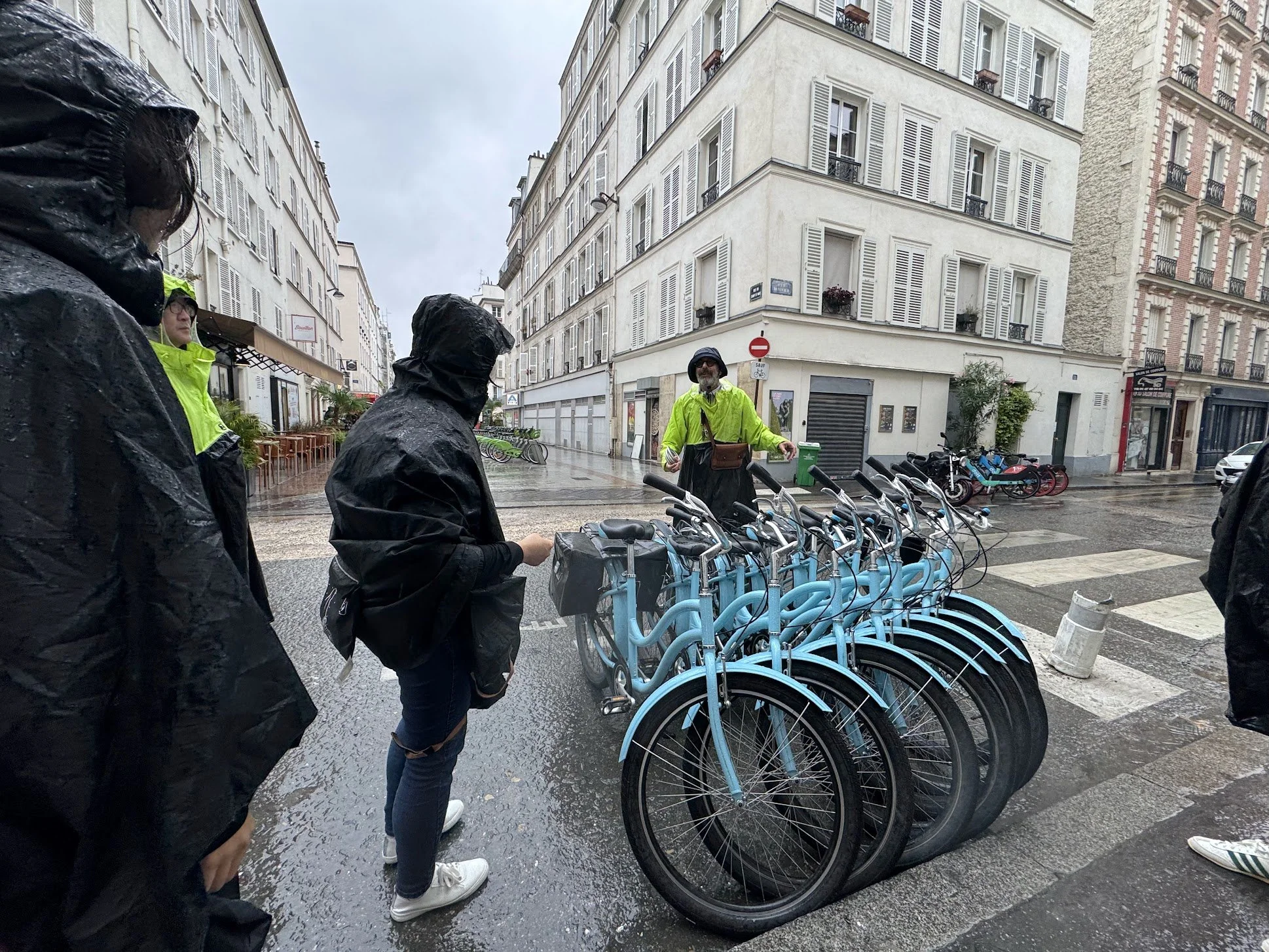 People wearing rain ponchos and a man with a helmet look at a row of blue rental bikes on a rainy city street with tall buildings.