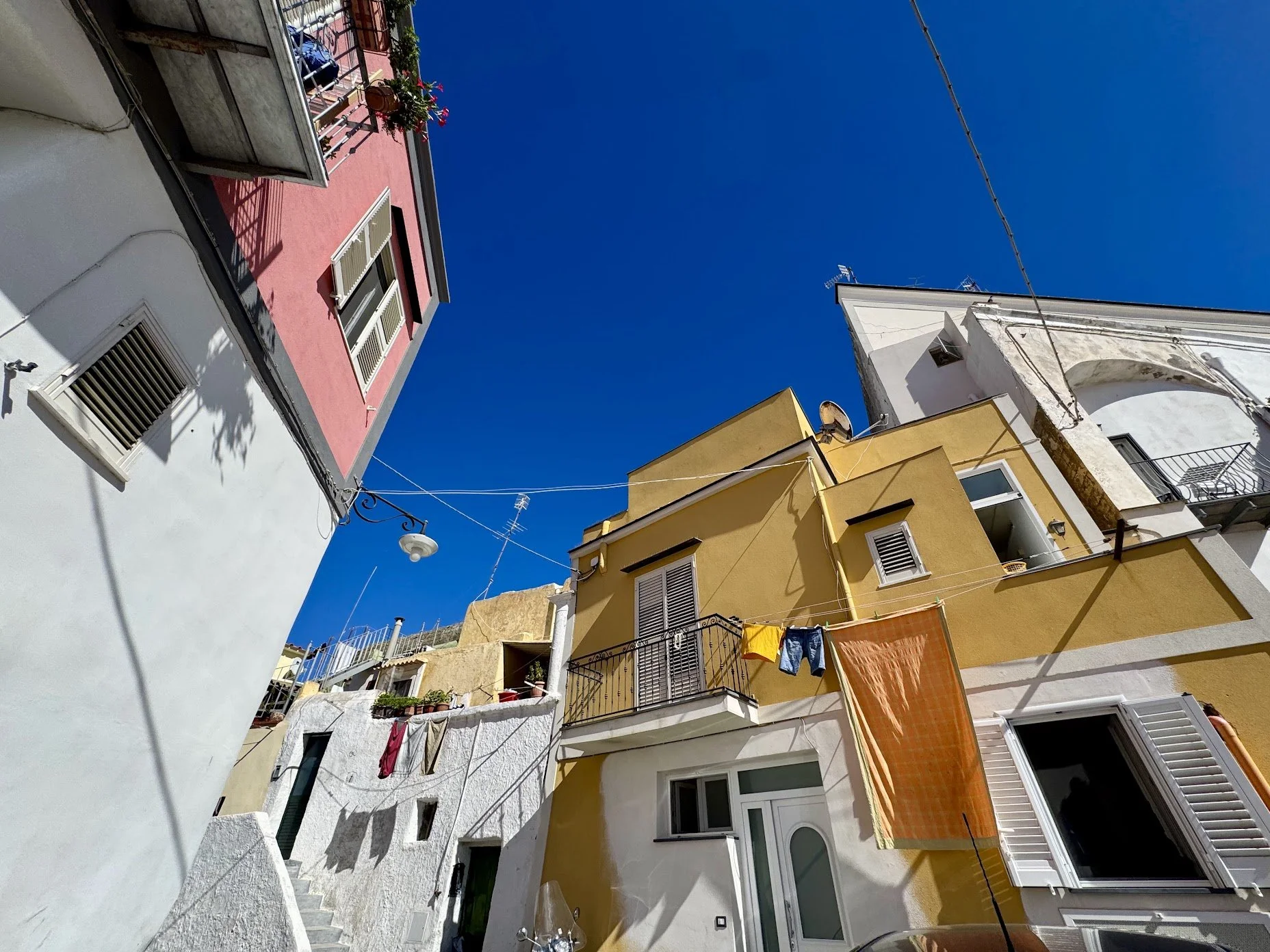 Colorful, multi-story buildings with balconies and laundry hanging across a bright blue sky in a Mediterranean seaside town.