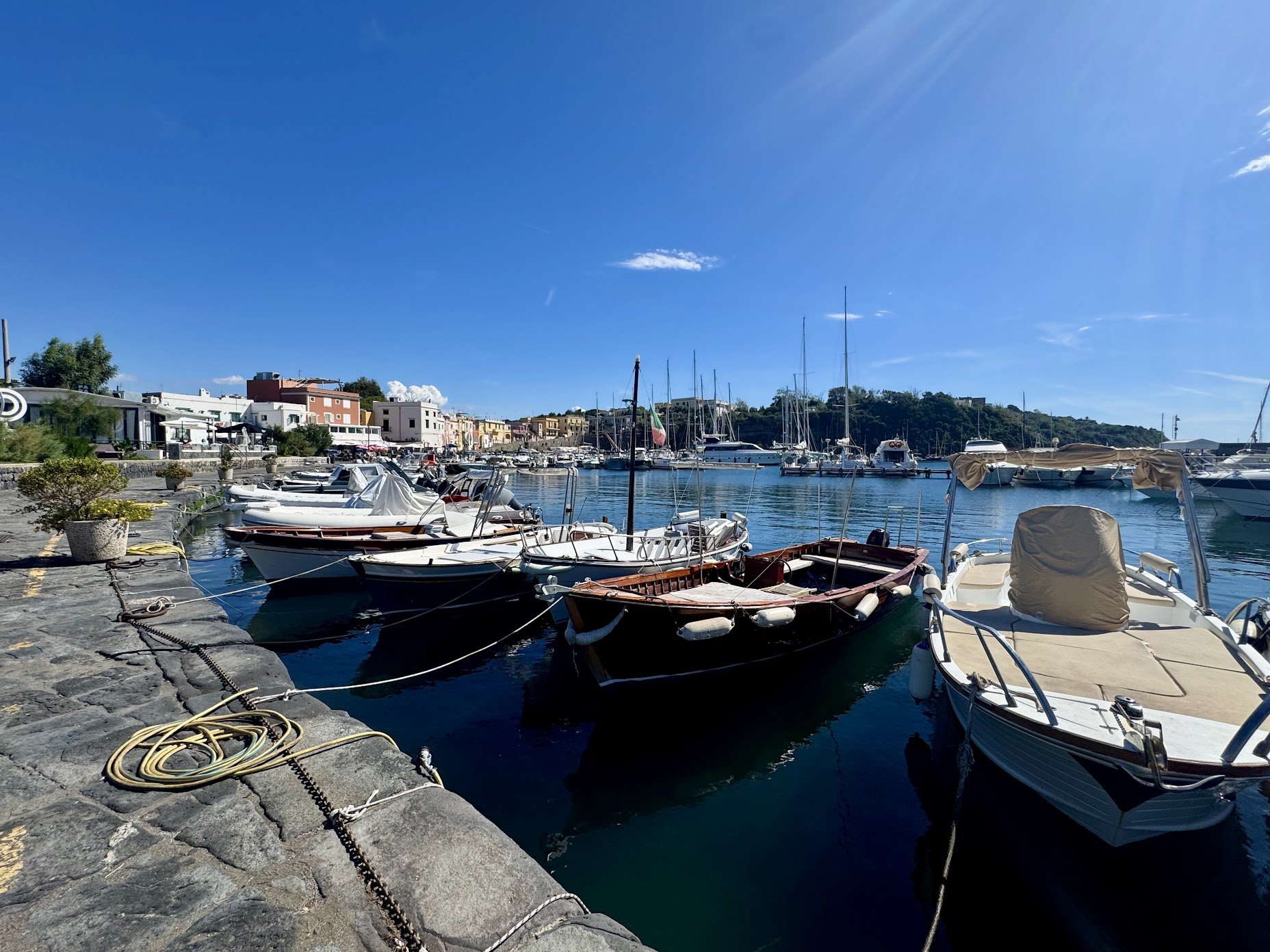 A marina with several boats docked along the pier under a clear blue sky, with colorful buildings lining the shore in the background.