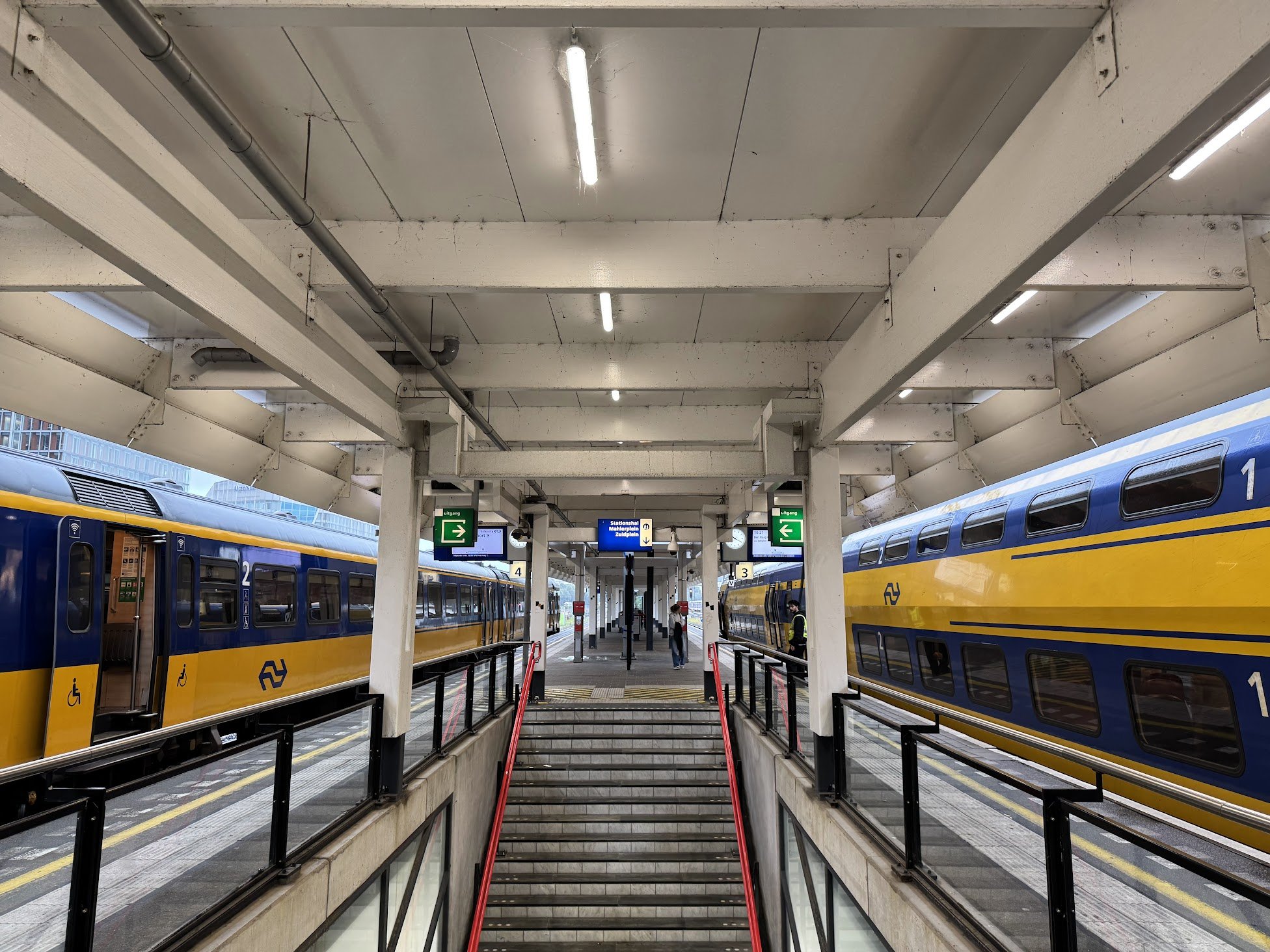Inside a train station with yellow and blue trains on both sides of a platform, with a staircase and escalator leading down. There are digital signs, people, and large windows in the background.