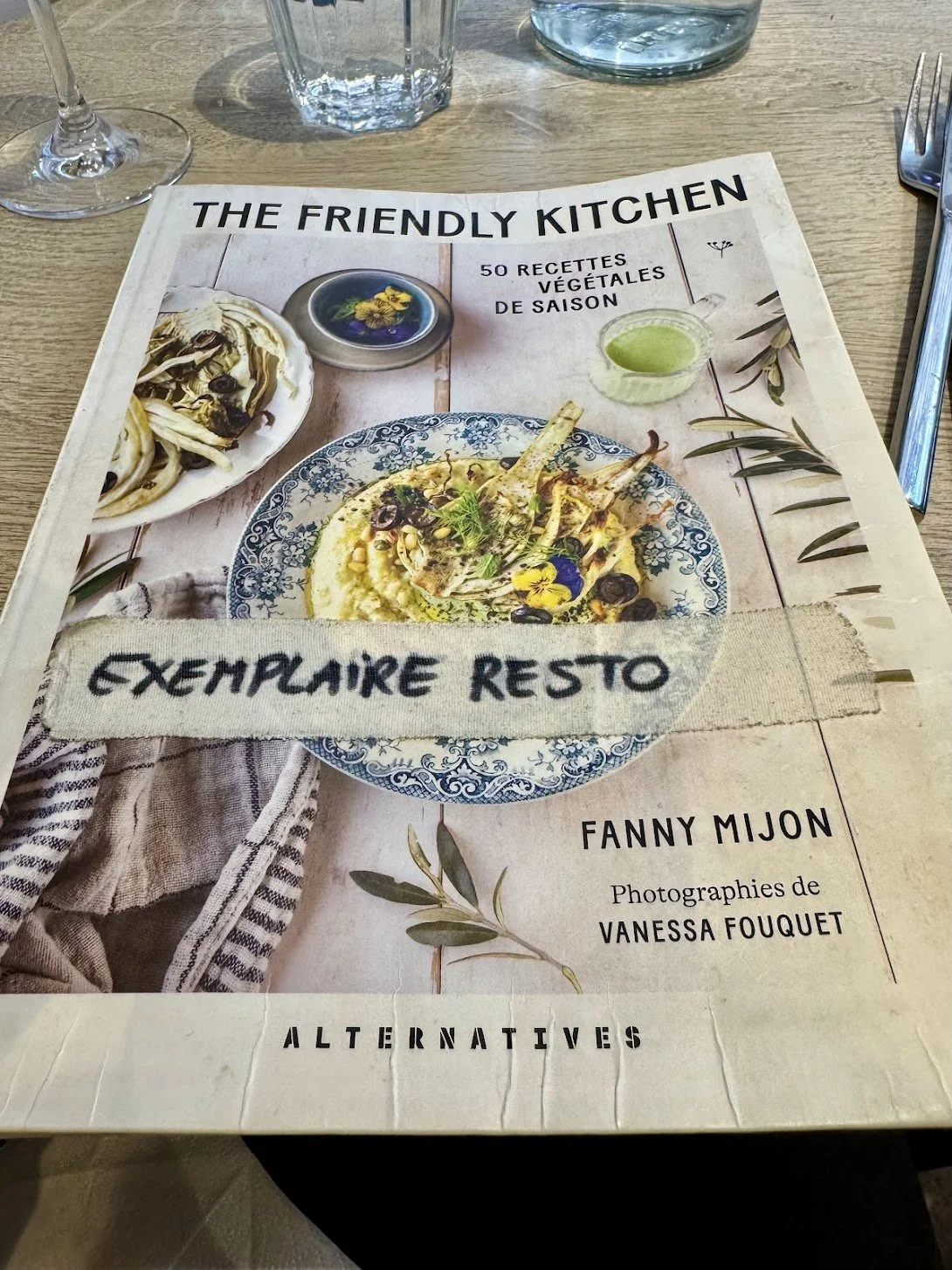A restaurant menu featuring vegetable recipes, with a focus on a dish of lamb chops with herbs, olives, and yellow sauce, garnished with edible flowers and herbs, accompanied by a side dish and drinks.