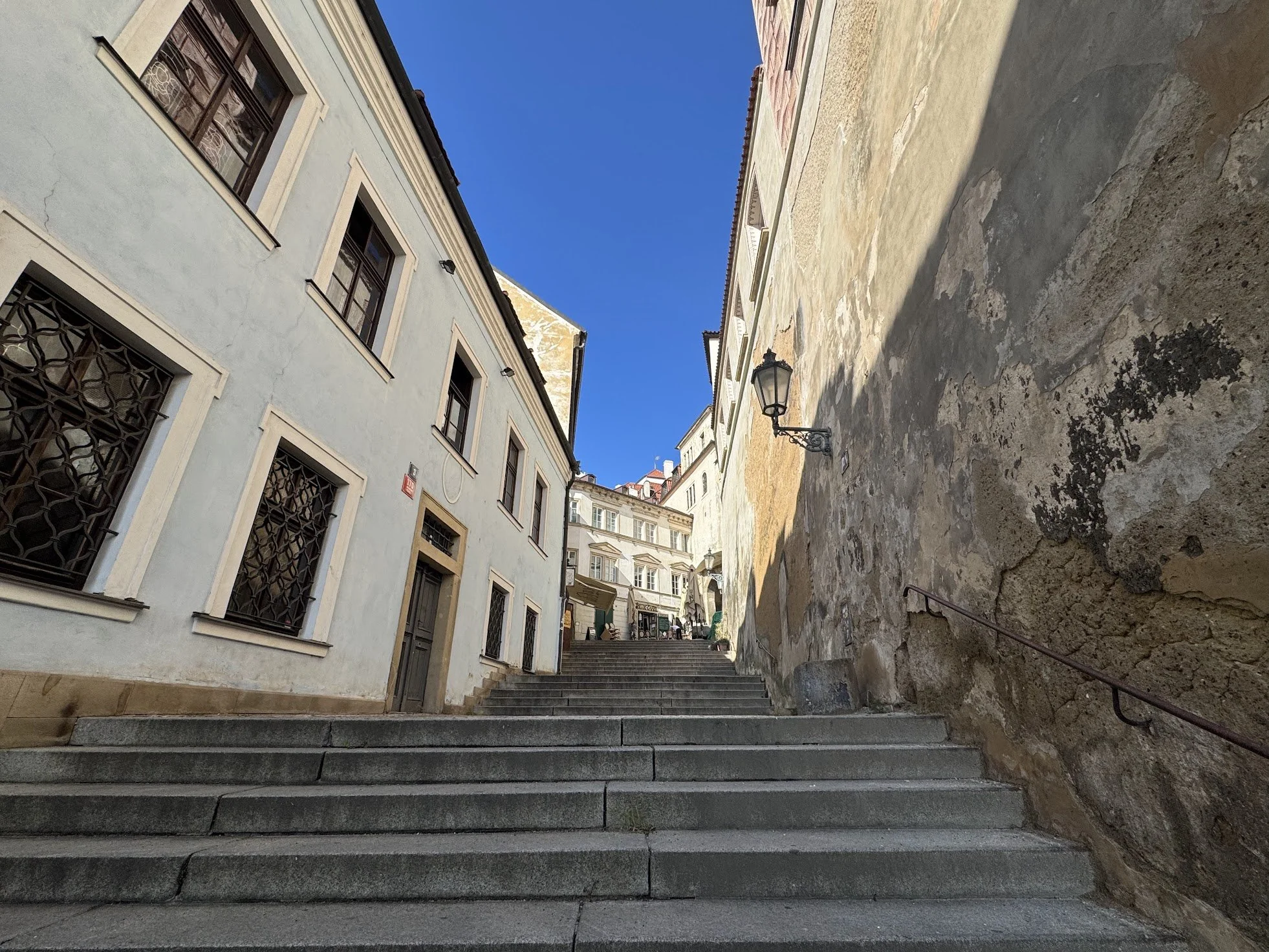 Stone steps leading upward between old buildings with European architecture, metal railings on the stairs, and a clear blue sky above.