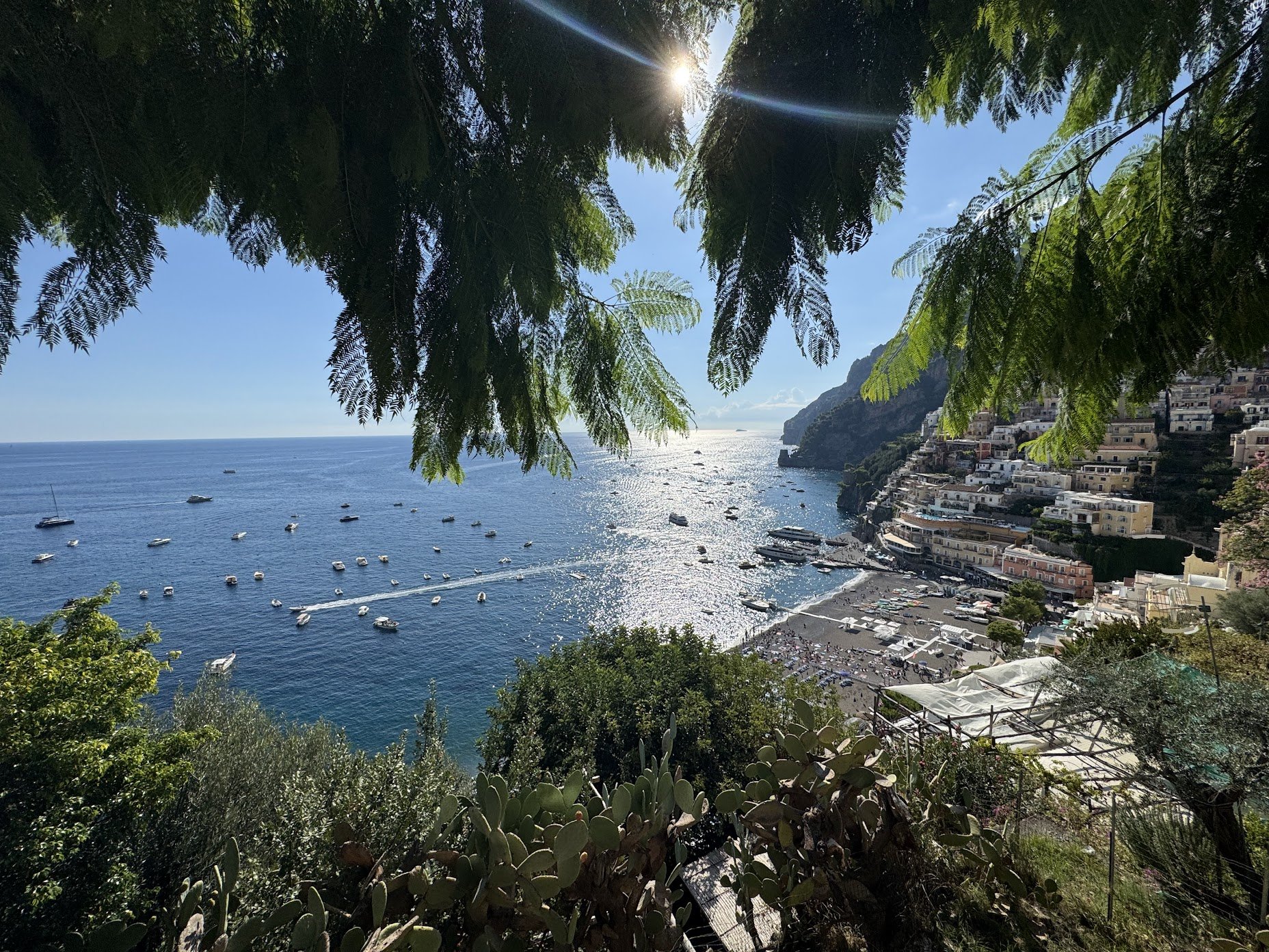 View of a coastal town with colorful buildings on a hillside, boats anchored in the water, and the sun shining through the trees.