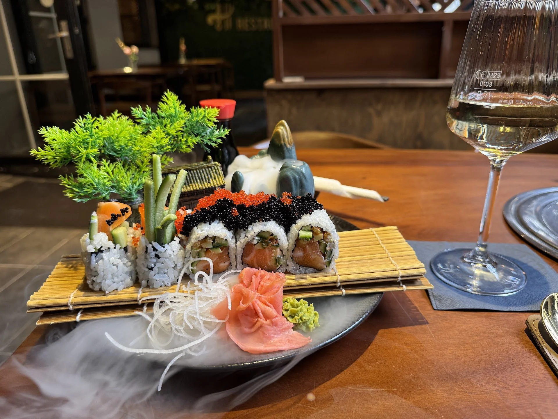 A plate of assorted sushi rolls with garnishes, a glass of water, and decorative plants on a wooden table.