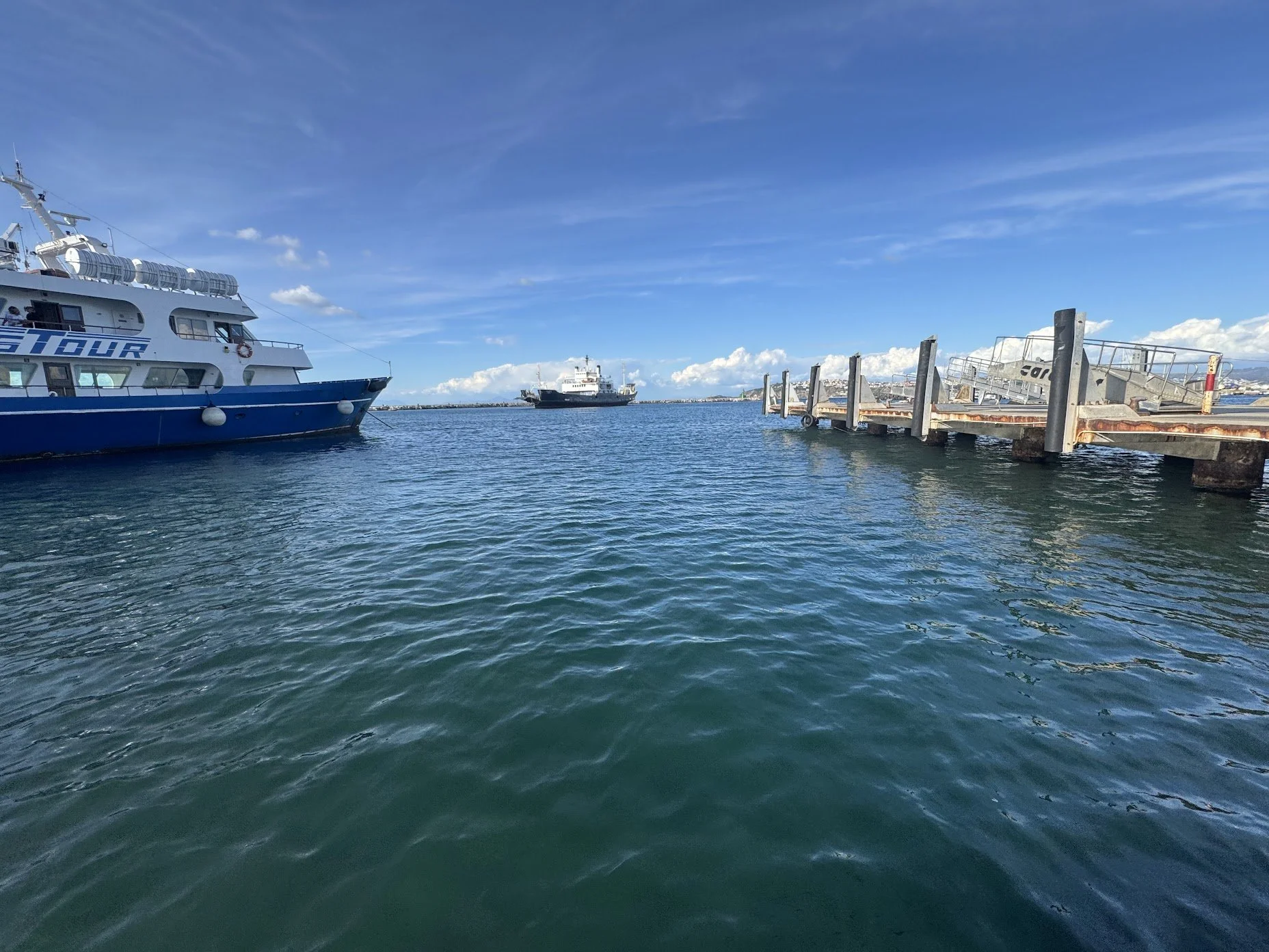 A marina with a large white and blue tour boat docked on the left, a small black and white vessel in the distance, and a weathered dock extending into the water on the right against a backdrop of a clear blue sky with some clouds.