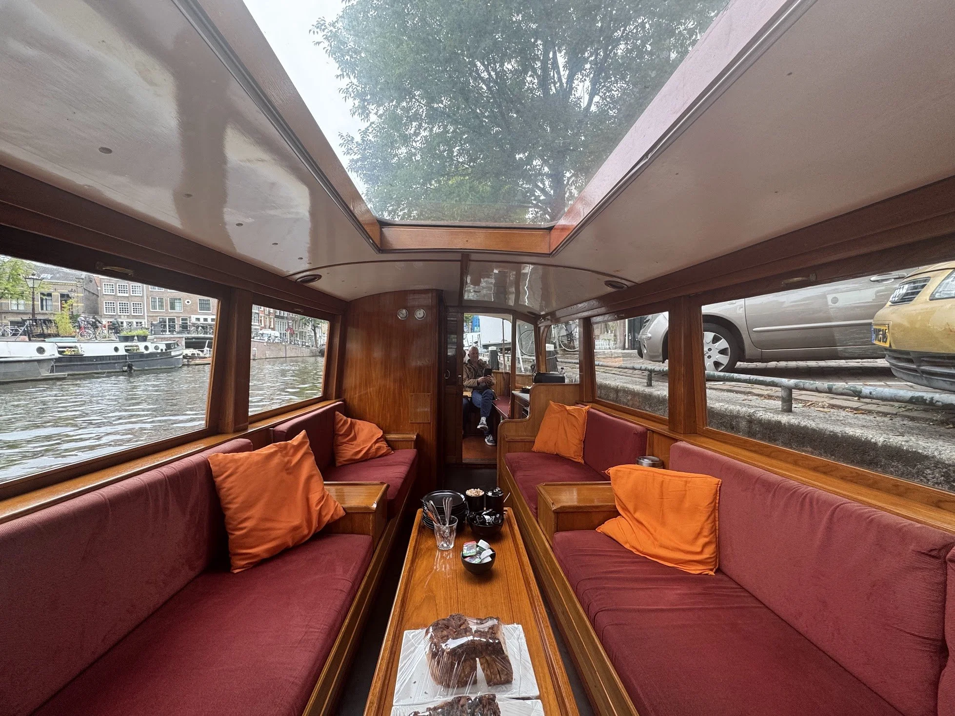 Interior of a boat with red cushioned seating and orange pillows, a wooden table with some food and drink, large windows showing boats and buildings on a canal, a person sitting in the background, and a large glass skylight with a tree visible outsid