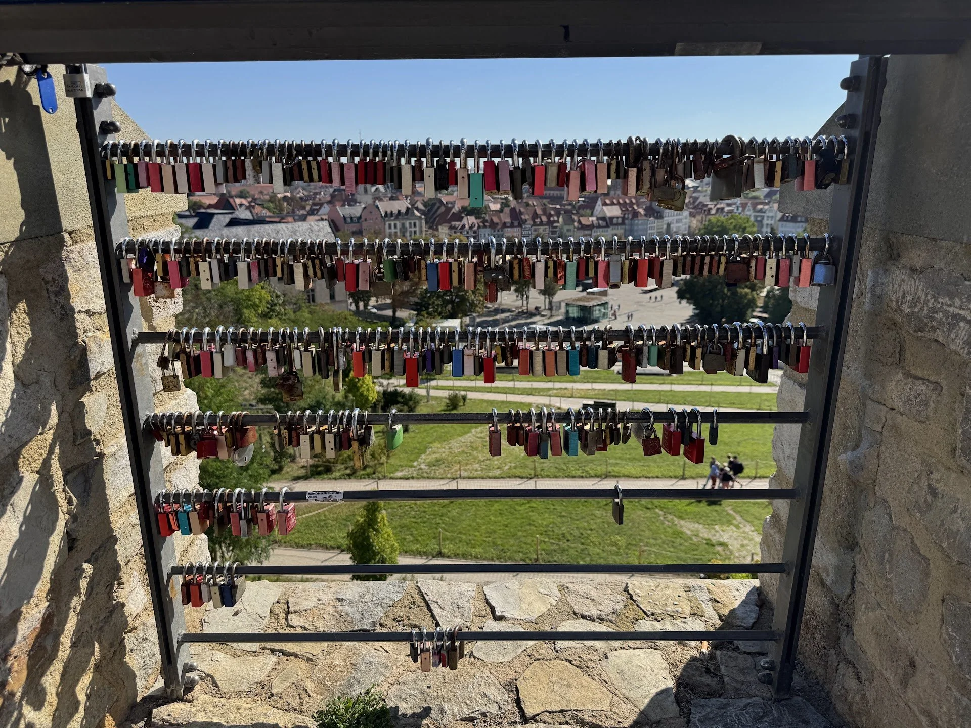 A metal rack filled with colorful padlocks attached to it, overlooking a town with houses and trees on a bright, sunny day.