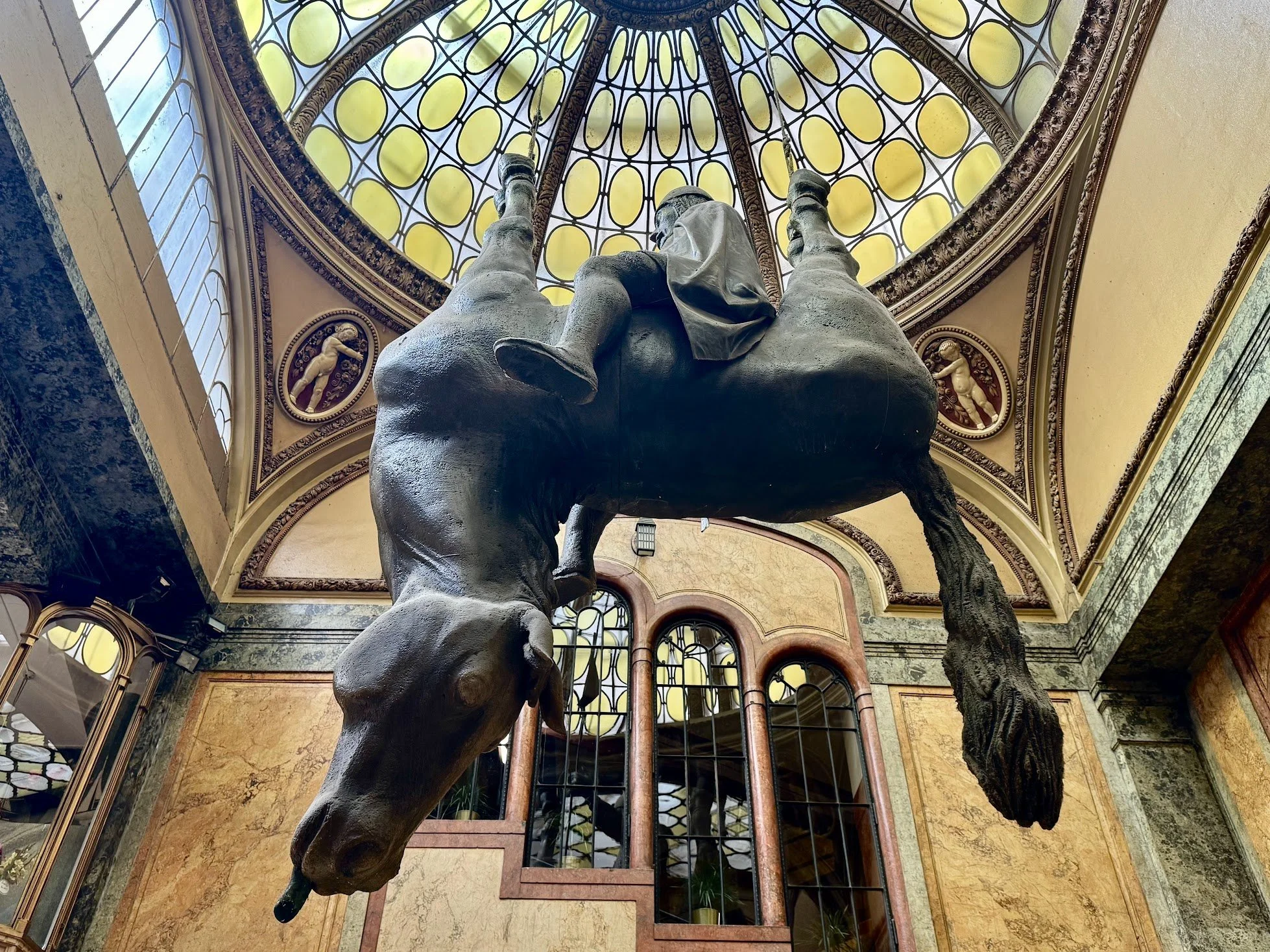 A bronze statue of an elephant with a person riding on its back, located inside a building with a glass and decorated ceiling.