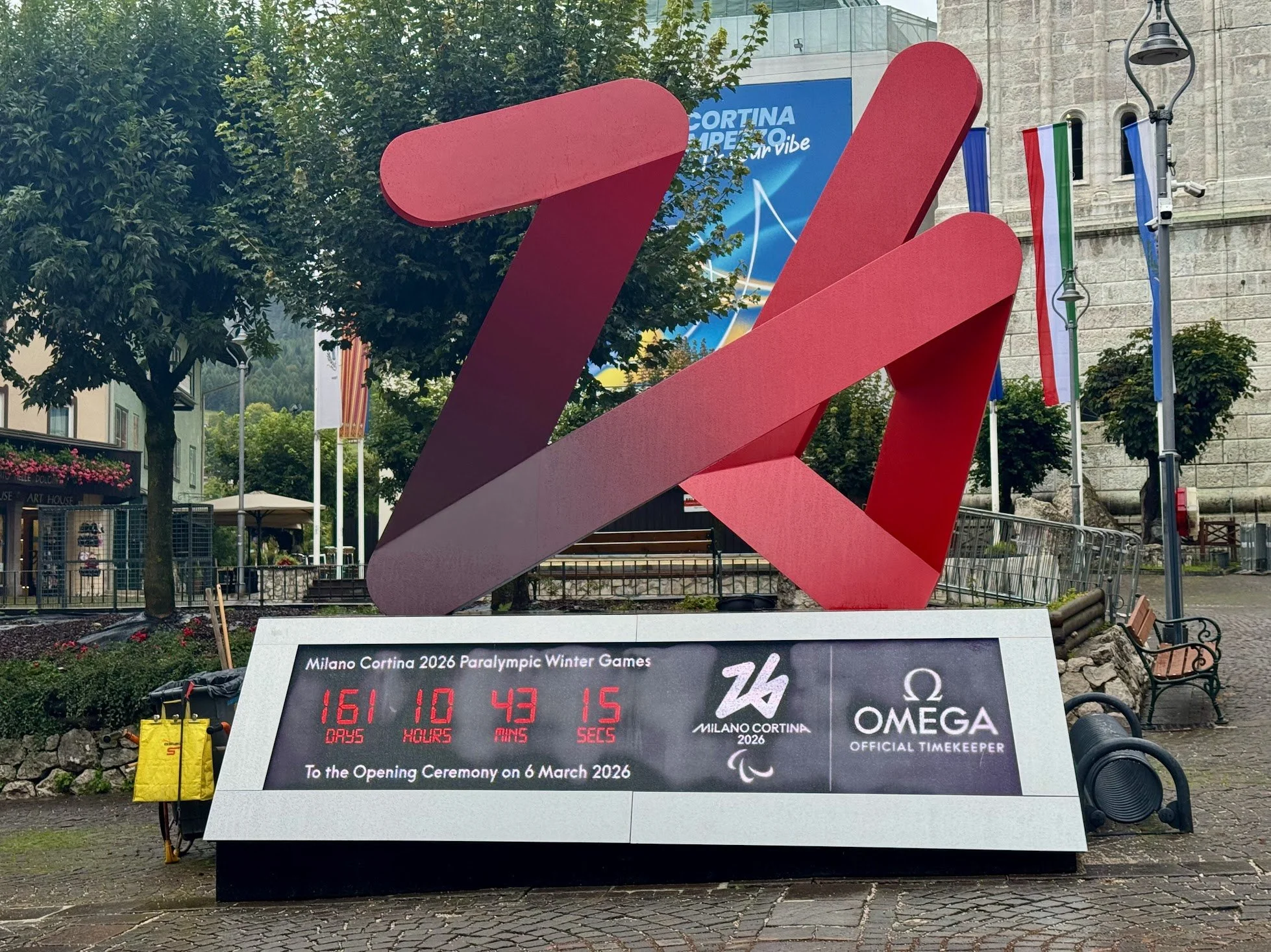 Red sculpture of a stylized, abstract figure in front of a building with trees and banners, supporting the 2026 Milan Cortina Paralympic Winter Games.