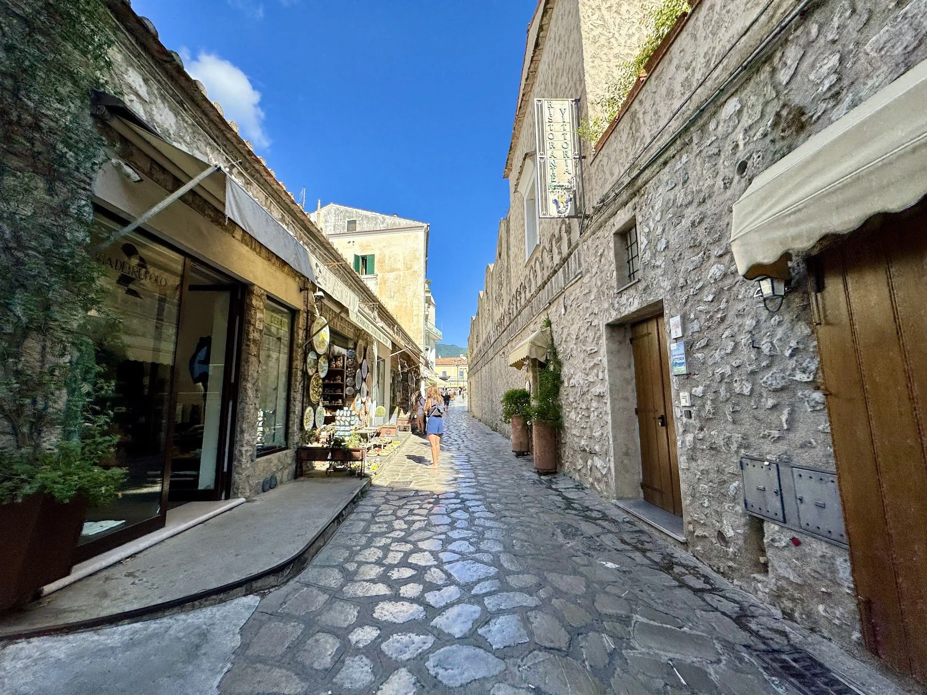 A narrow cobblestone street lined with stone buildings, shops, and an outdoor market on a sunny day