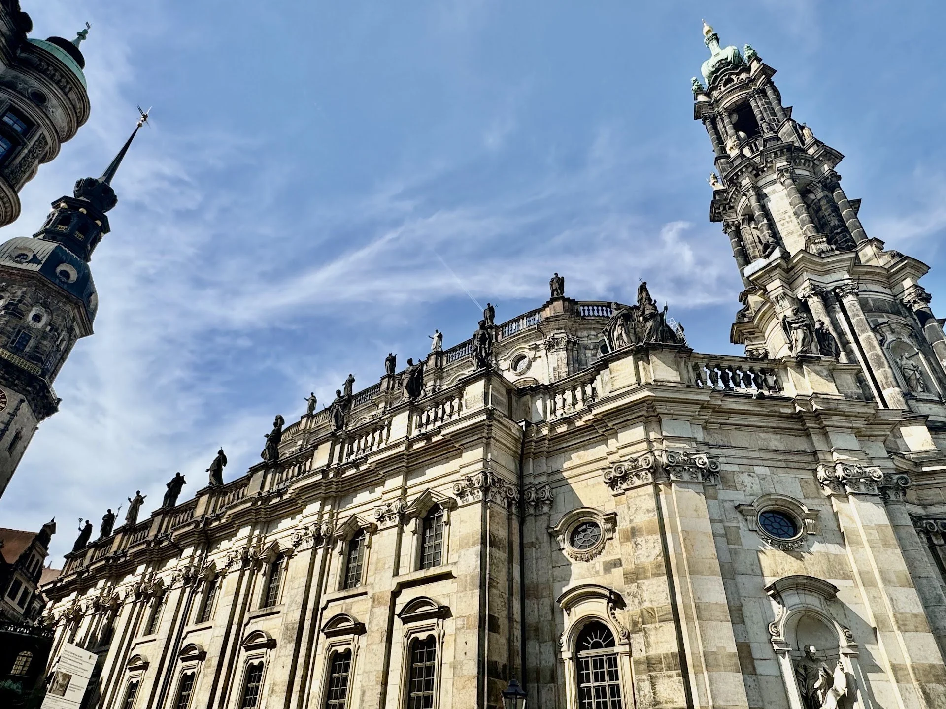 A historic European cathedral with detailed stone architecture and statues, topped with multiple spires and a green-domed tower, under a partly cloudy blue sky.