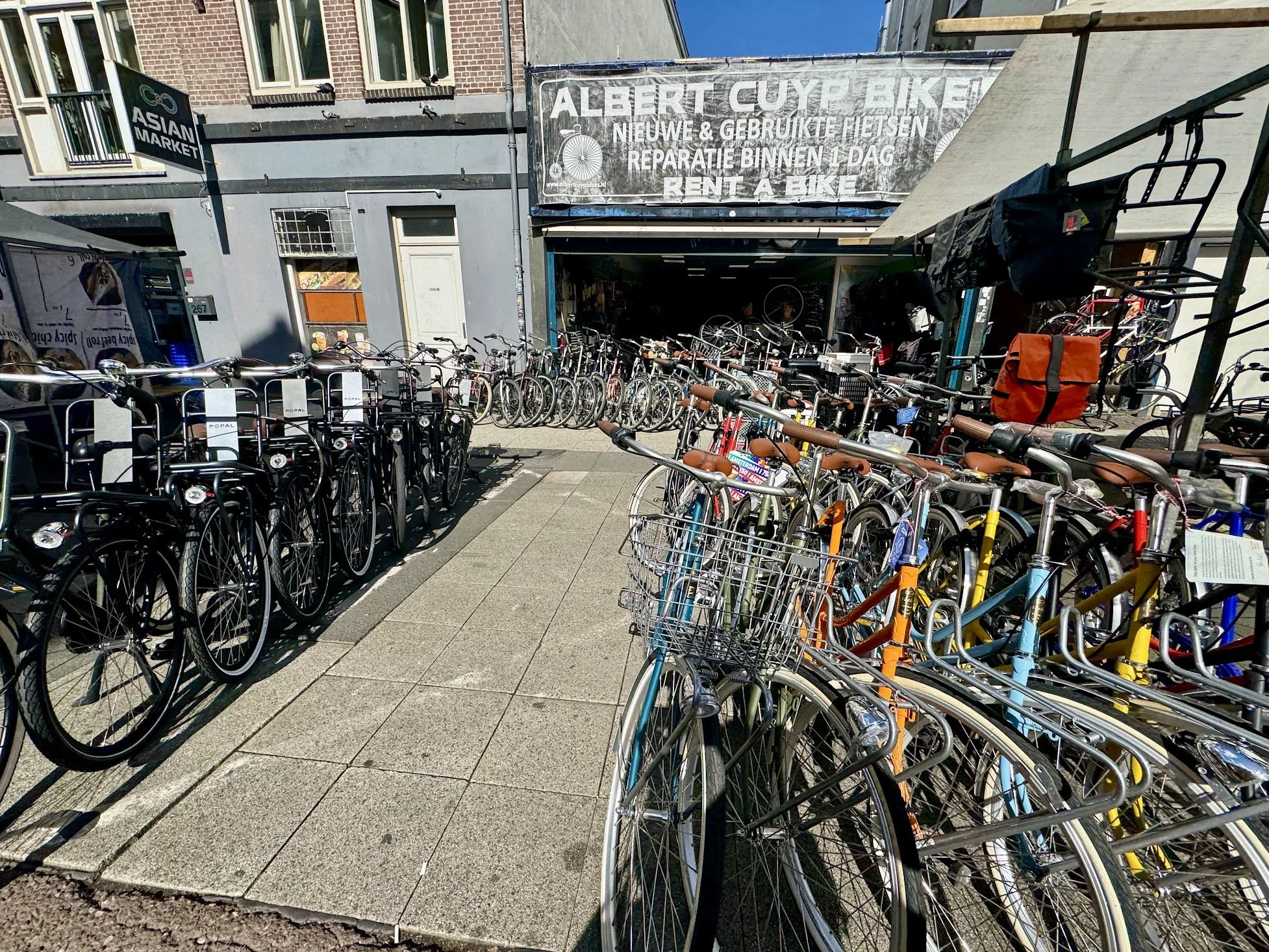 Bike rental shop front with numerous bicycles parked outside, sign for 'Asian Market' on left, and shop sign reading 'Albert Cuypmarkt' indicating a market that rents bikes and repairs bikes, located in an urban area with brick buildings.