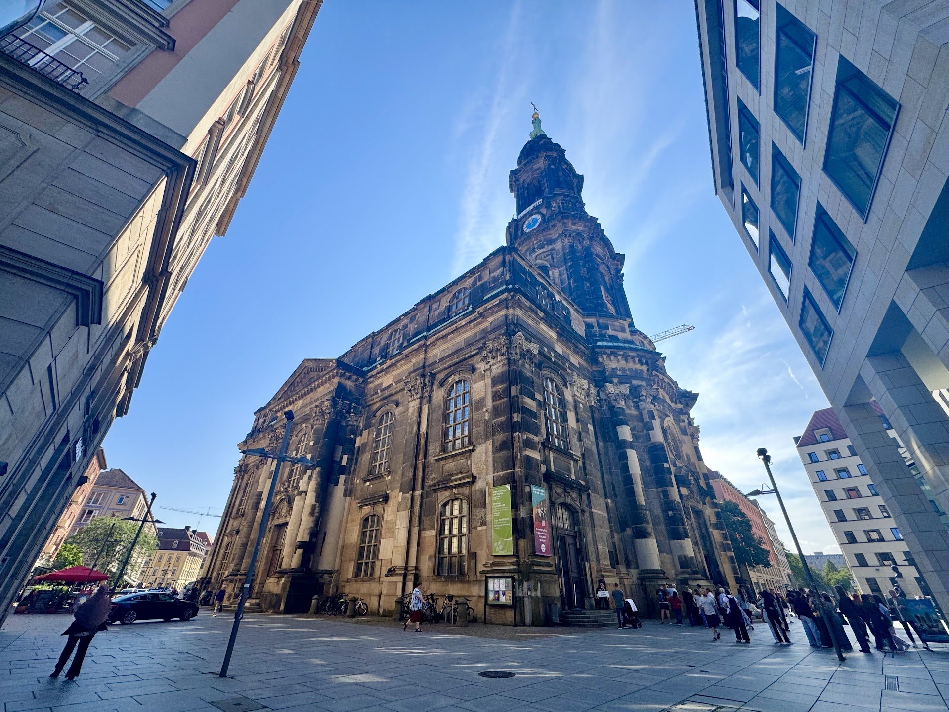 A historic church building with ornate architecture, tall windows, and a clock on its front facade, surrounded by modern high-rise buildings, with people walking in the street and a clear blue sky overhead.