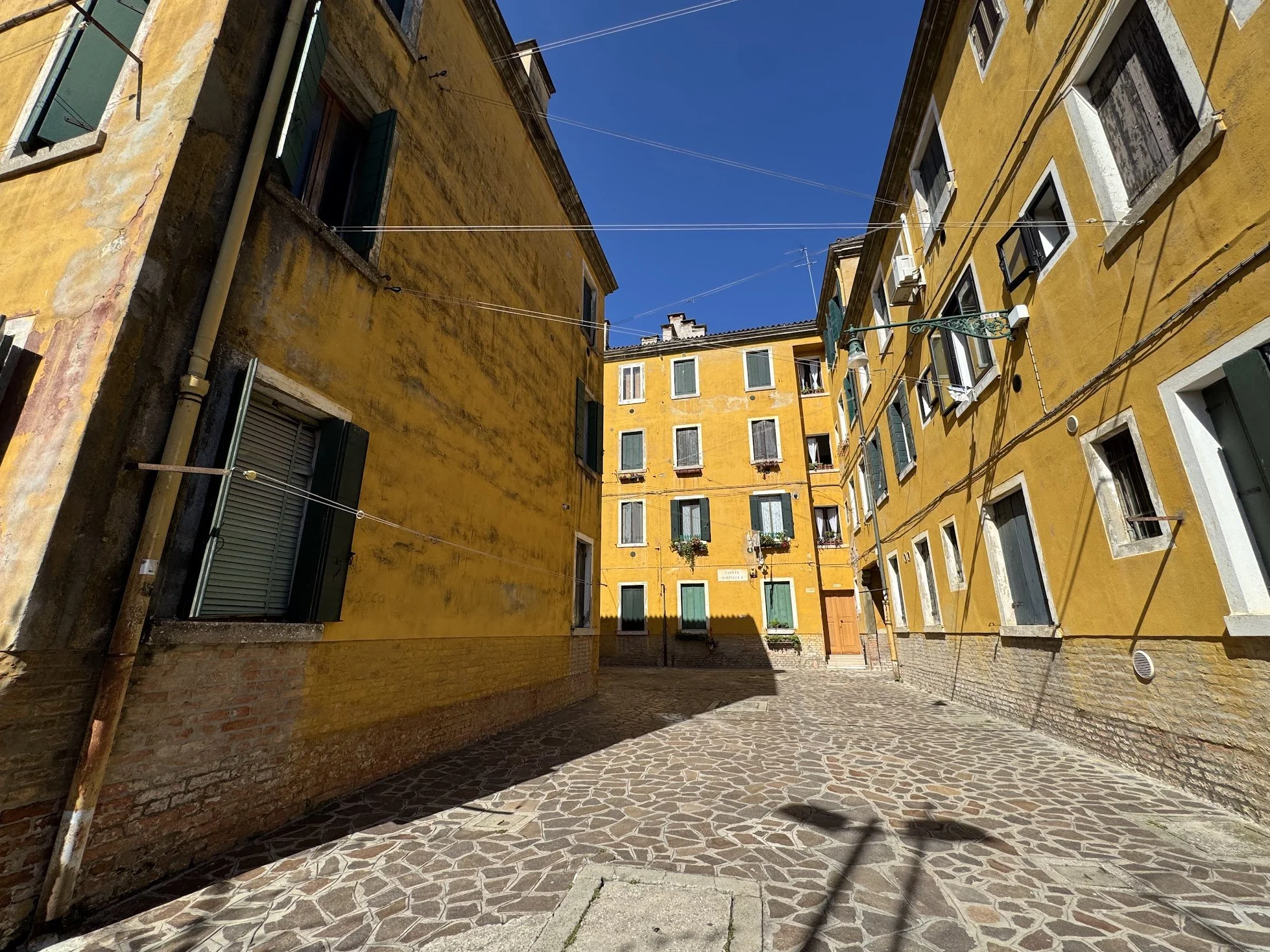 A narrow European alleyway with cobblestone pavement, yellow buildings with green shutters, and blue sky overhead.