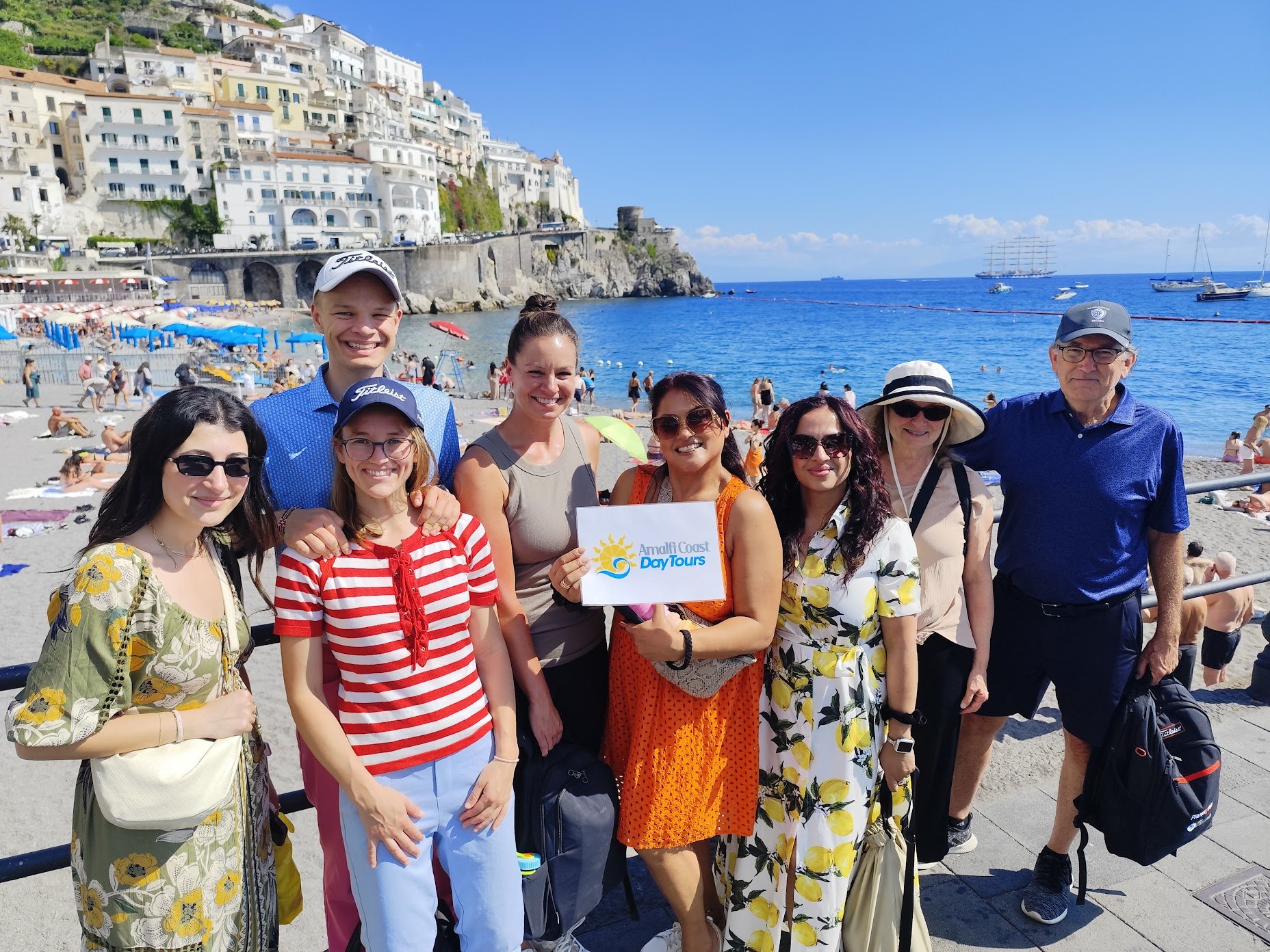 A group of seven people standing together on a beach, smiling at the camera, with the ocean, boats, and colorful buildings on a hillside in the background. One person is holding a sign that says "Ammilf Coast Day Tours."