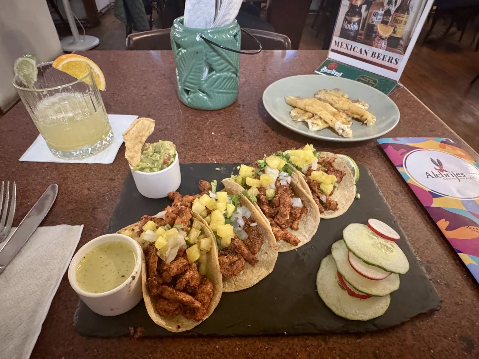 A table at a Mexican restaurant featuring tacos topped with pineapple, guacamole with a chip, cucumber and radish slices, and two cups of green salsa, with a drink containing lime and orange slices and a menu promoting Mexican beers.