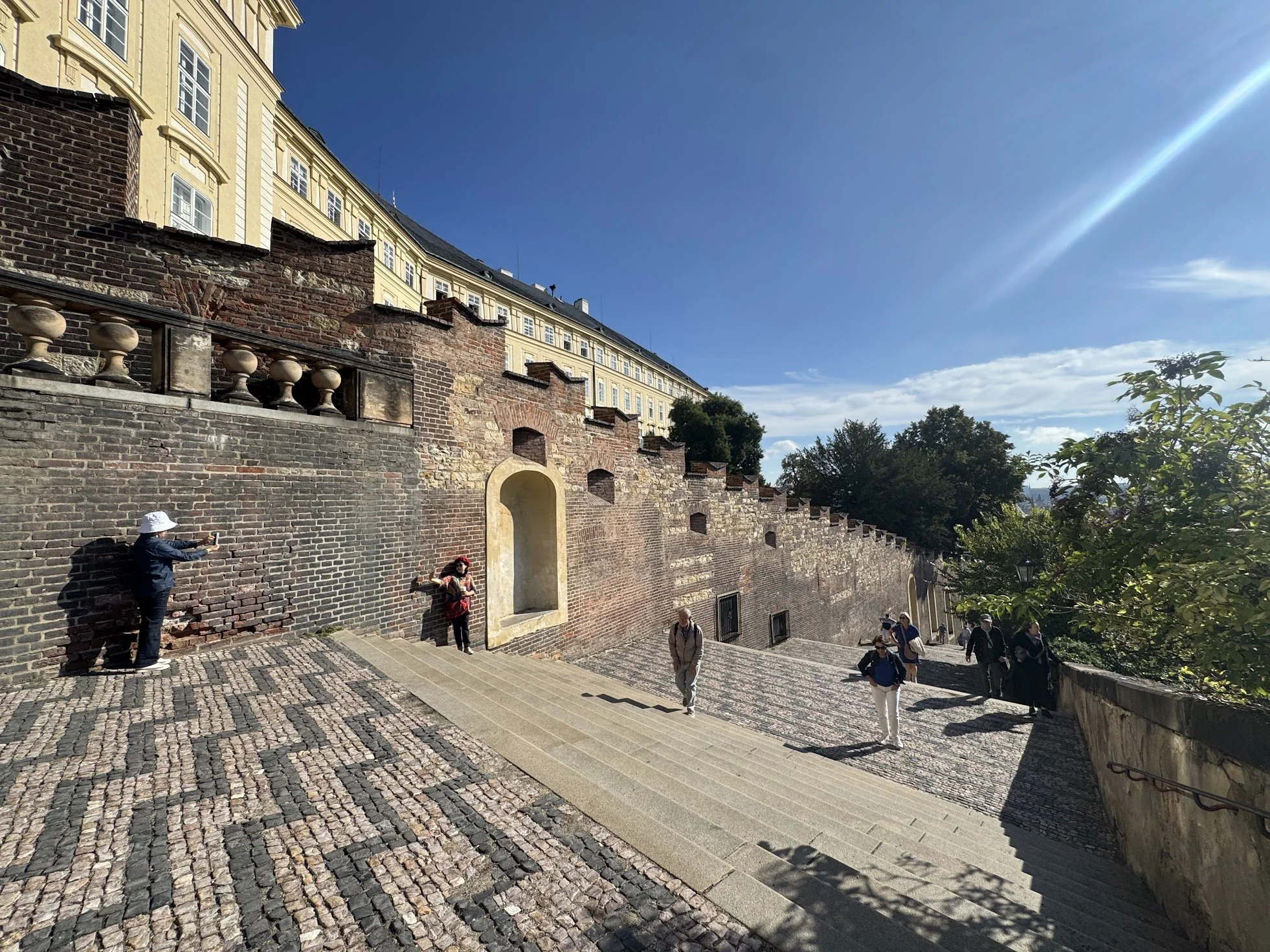 People walking along a cobblestone path beside a historic brick wall, with a large yellow building with many windows above it, under a clear blue sky with sunlight streaming in from the right.