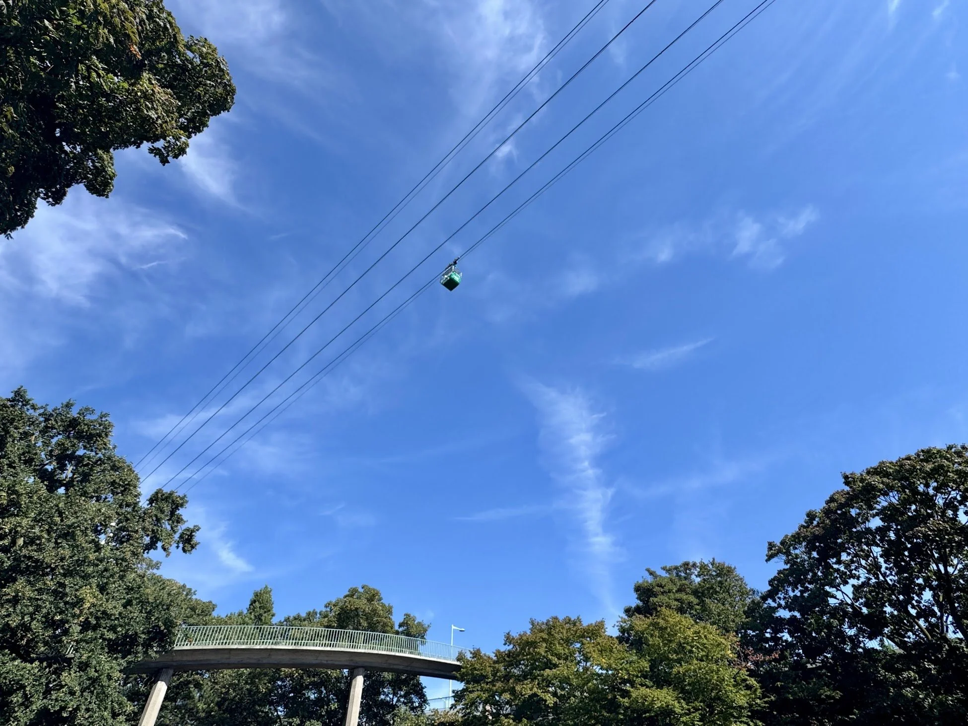 Blue sky with wispy clouds, trees on the lower part, and a gondola lift cable crossing from left to right, with a green gondola hanging from the cable.