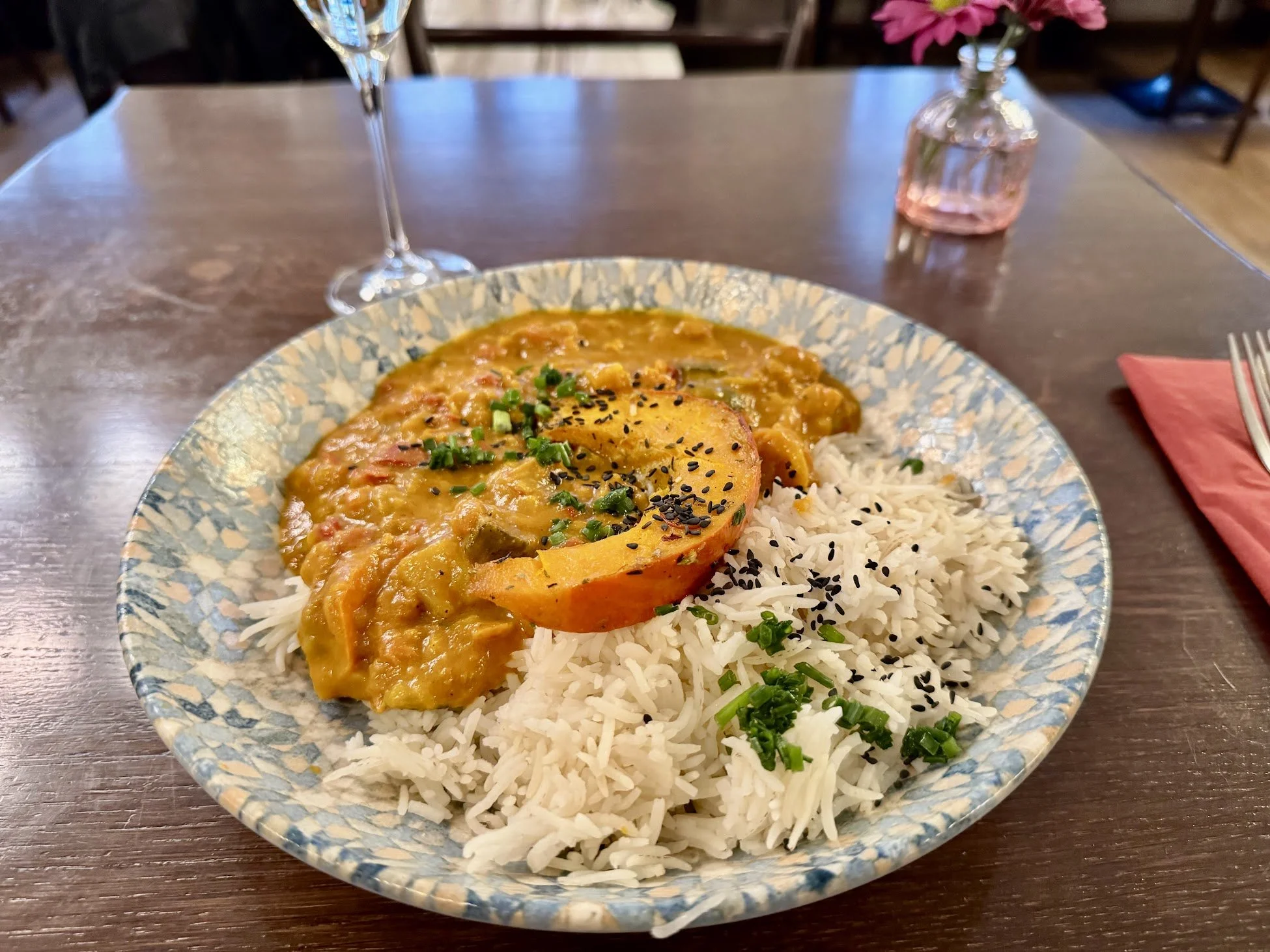 A plate of rice with black sesame seeds and green onions, topped with a slice of stuffed pumpkin in a yellow sauce, on a wooden table with a wine glass, a pink napkin, and a small pink flower vase in the background.