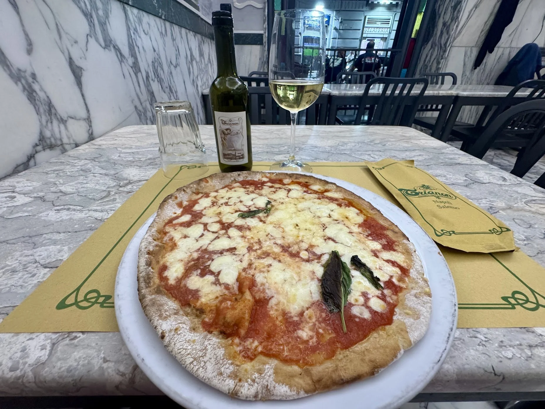 A pizza with cheese and basil leaves on a white plate, placed on a yellow paper mat on a marble table. Behind, there is a bottle of white wine, a glass of wine, and an upside-down glass in a restaurant setting with chairs and a marble wall.