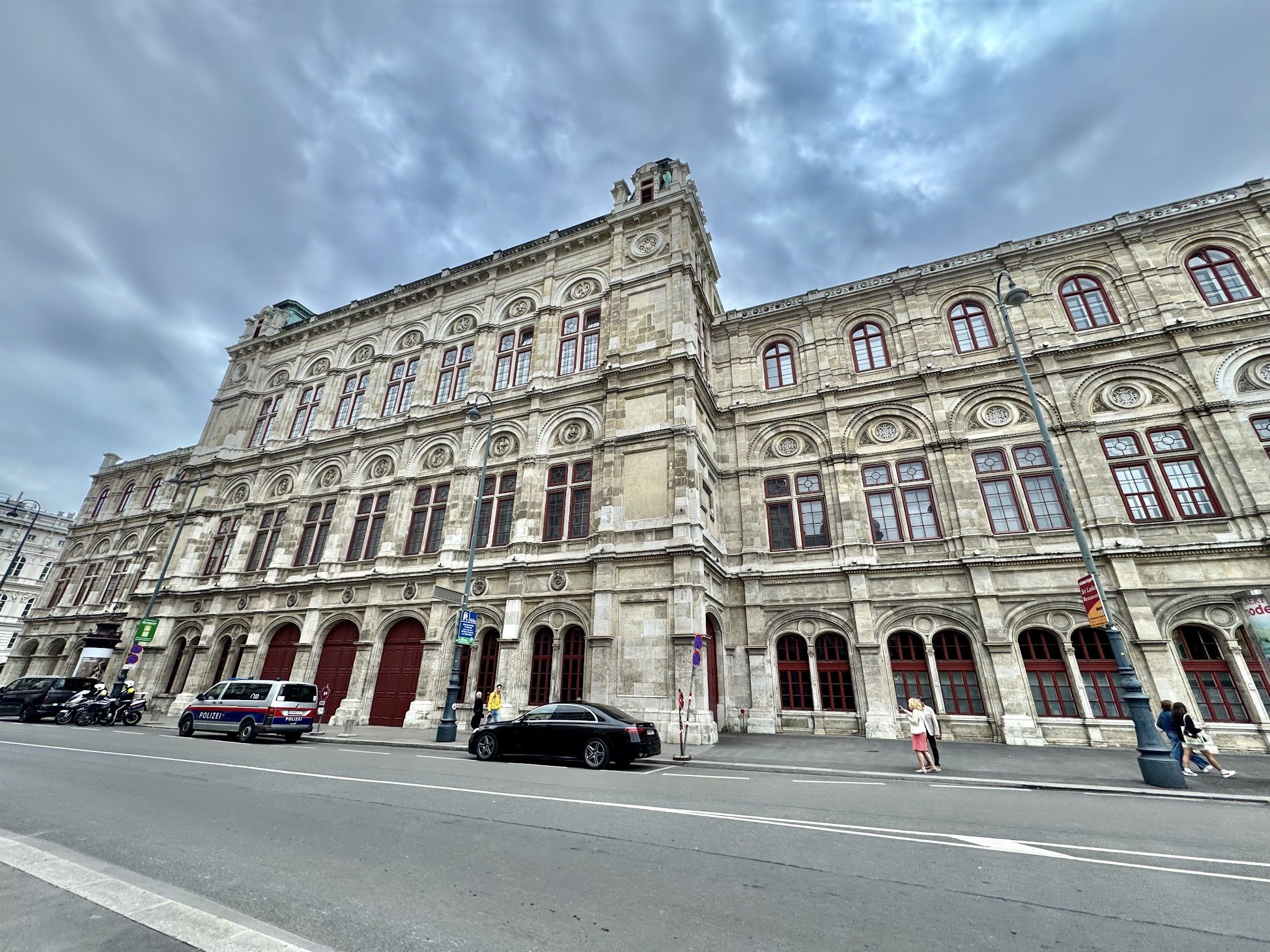 A large historic building with detailed stone architecture and red window frames, situated on a city street. Several cars and motorcycles are parked in front, with a police vehicle visible. Pedestrians are walking along the sidewalk under street lamp